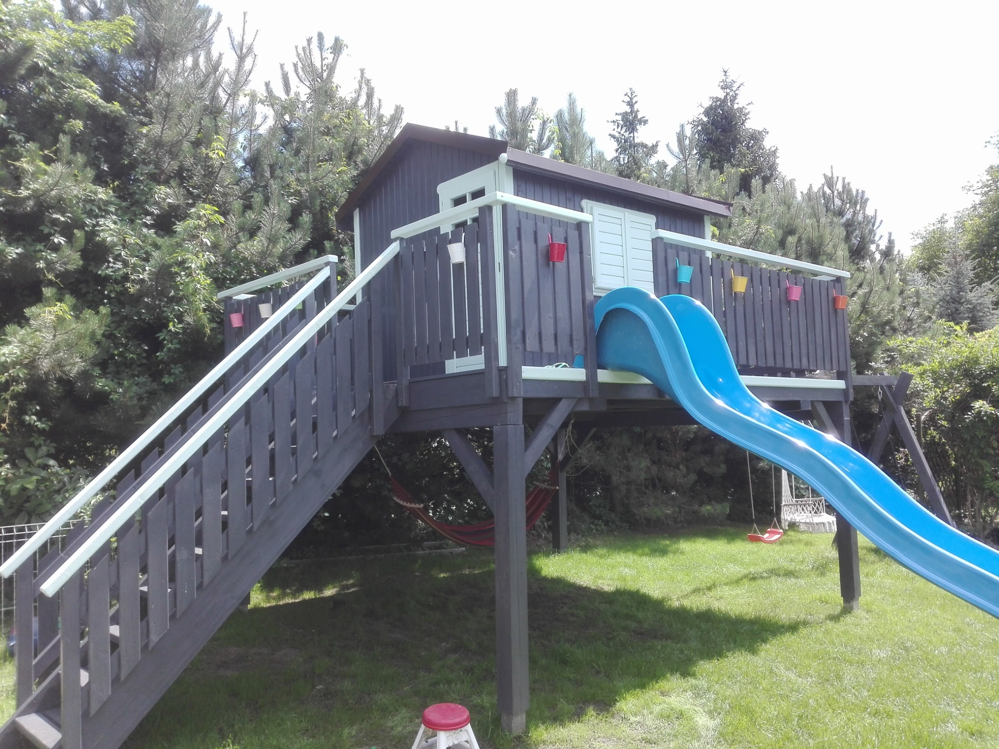 Drewniany domek dla dzieci na podwyższeniu z niebieską zjeżdżalnią, schodami z balustradą i wiszącymi kolorowymi doniczkami, otoczony zielenią.