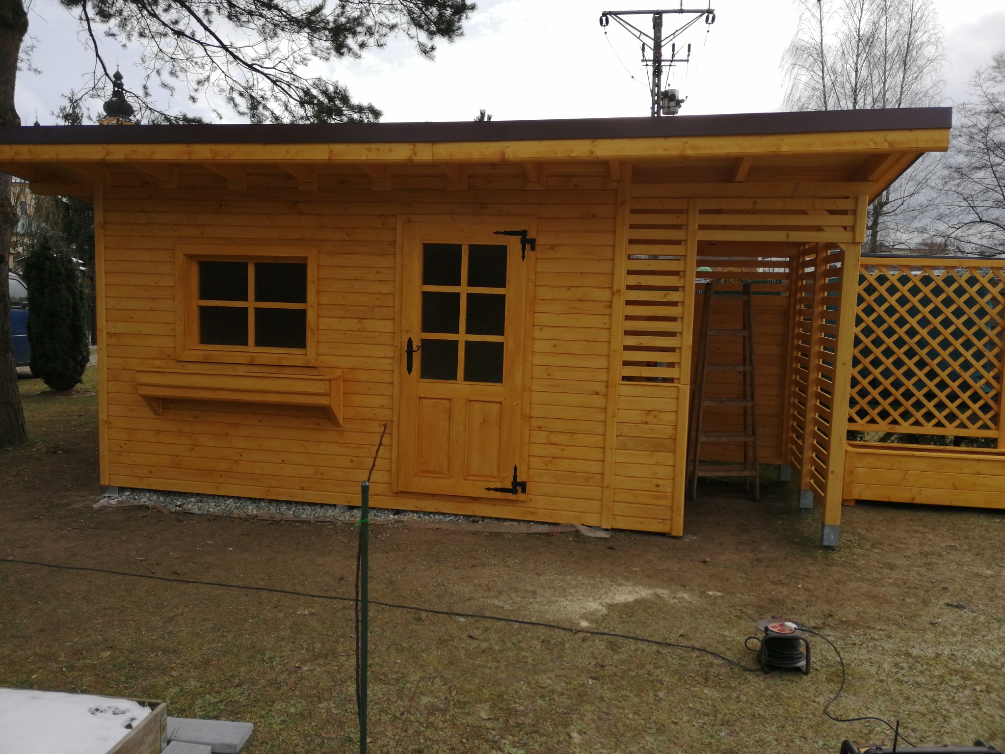 Nowa, jasna, drewniana konstrukcja z oknem, drzwiami i ażurową dobudówką, prawdopodobnie w trakcie montażu, widoczny zwinięty kabel elektryczny na ziemi.