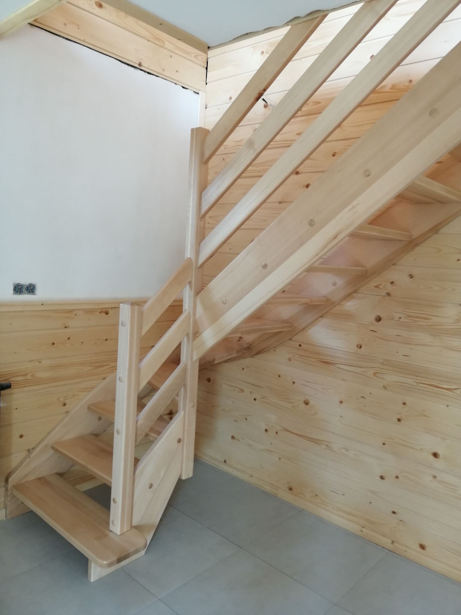 Jasne, drewniane schody z balustradą wewnątrz domu, z widoczną boazerią i szarą podłogą.