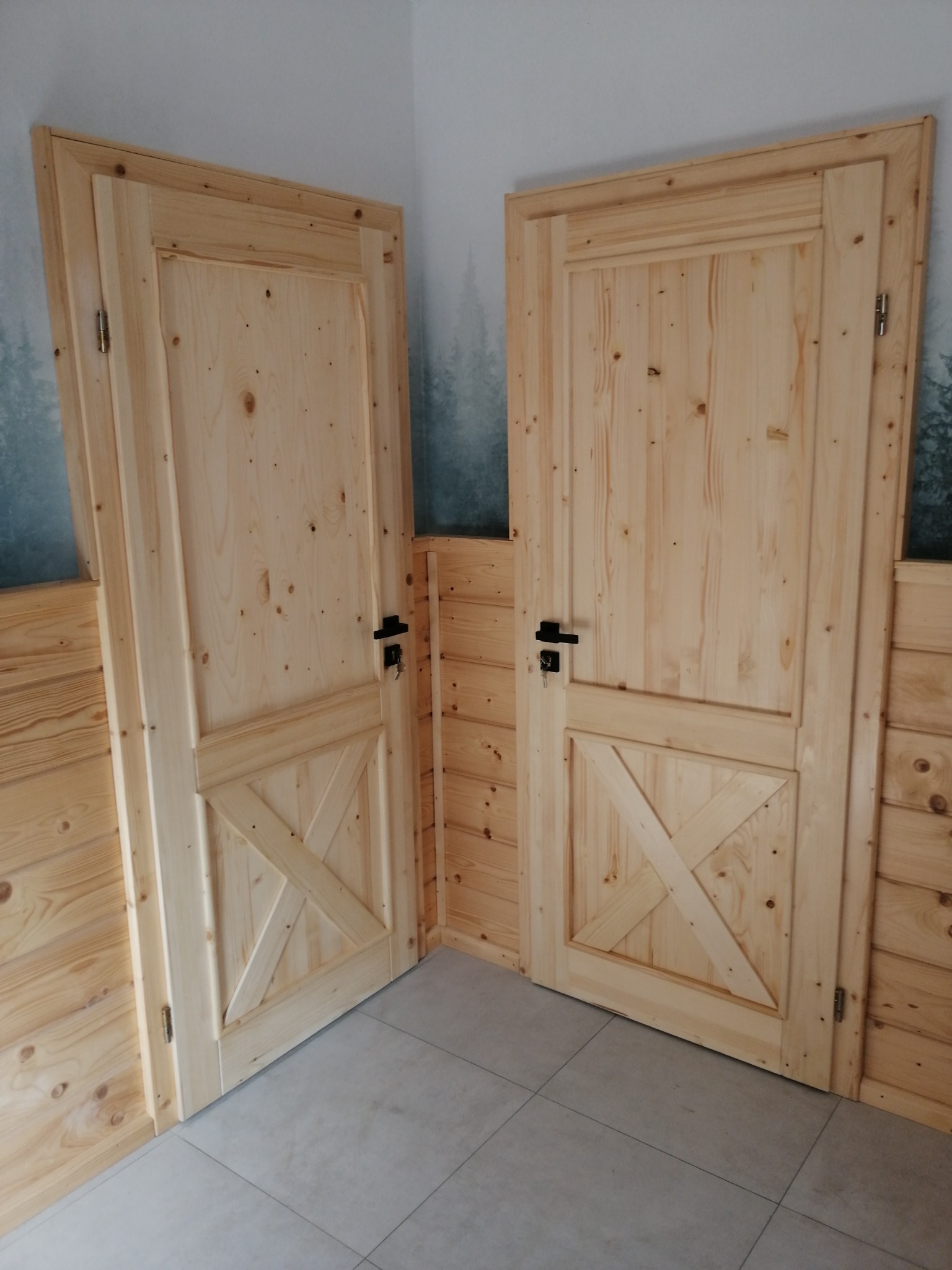 Dwie sosnowe, surowe drzwi wewnętrzne z czarnymi klamkami, wykończone krzyżowym wzorem, zamontowane w pomieszczeniu z boazerią i szarą podłogą.