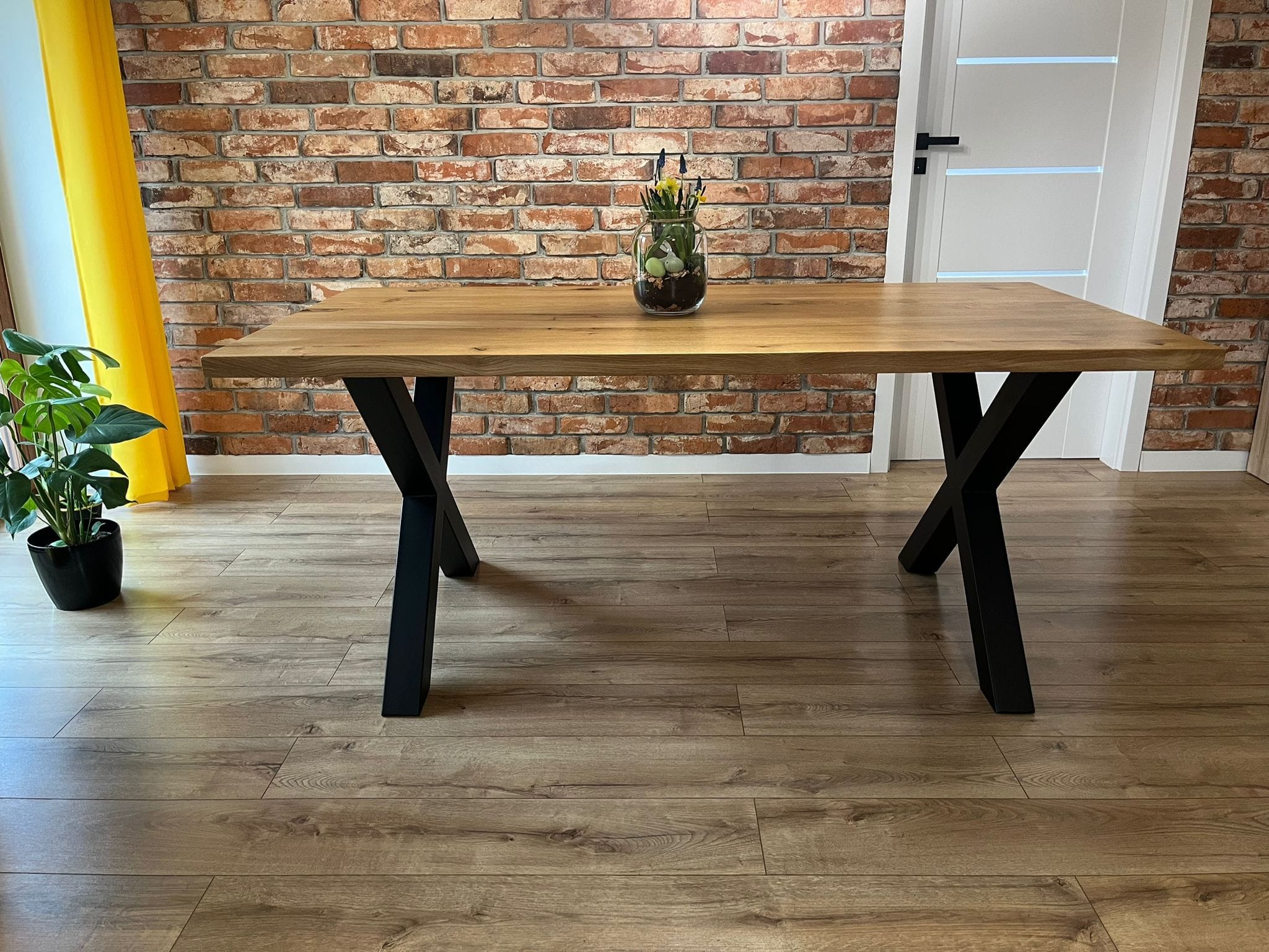 Stół z blatem z jasnego drewna na czarnych, metalowych nogach w kształcie litery X, ustawiony na podłodze z paneli imitujących drewno, z ceglaną ścianą w tle i dekoracją w szklanym wazonie.