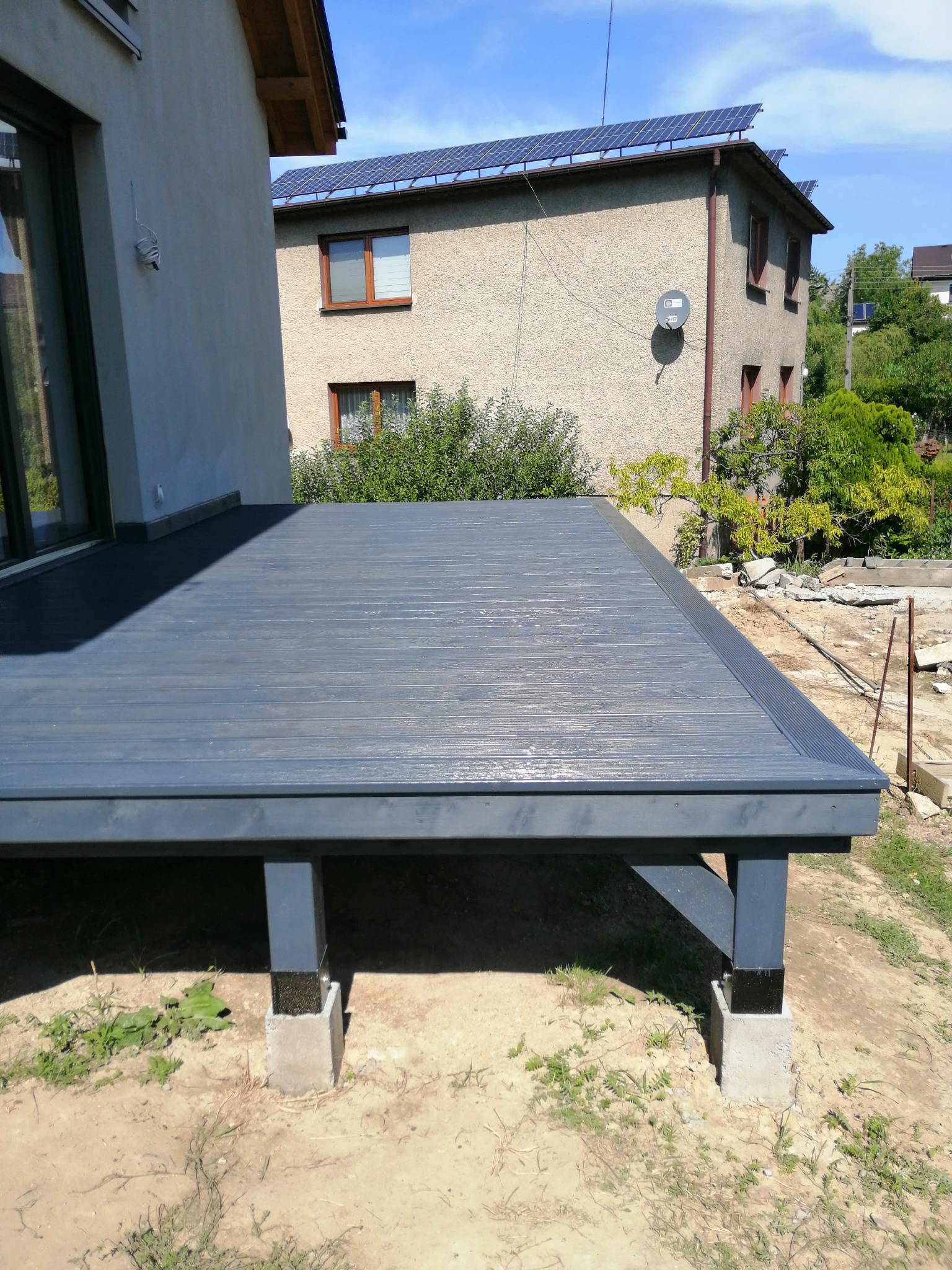 Nisko położony, pomalowany na szaro taras z drewna, wsparty na betonowych fundamentach, widok na dom z panelami słonecznymi w tle.