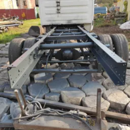 Surowa rama podwozia samochodu ciężarowego, pomalowana na szaro, stoi na nierównym podłożu z betonowych płyt. Widoczne opony, osie i elementy zawieszenia. W tle budynek i sterta złomu.