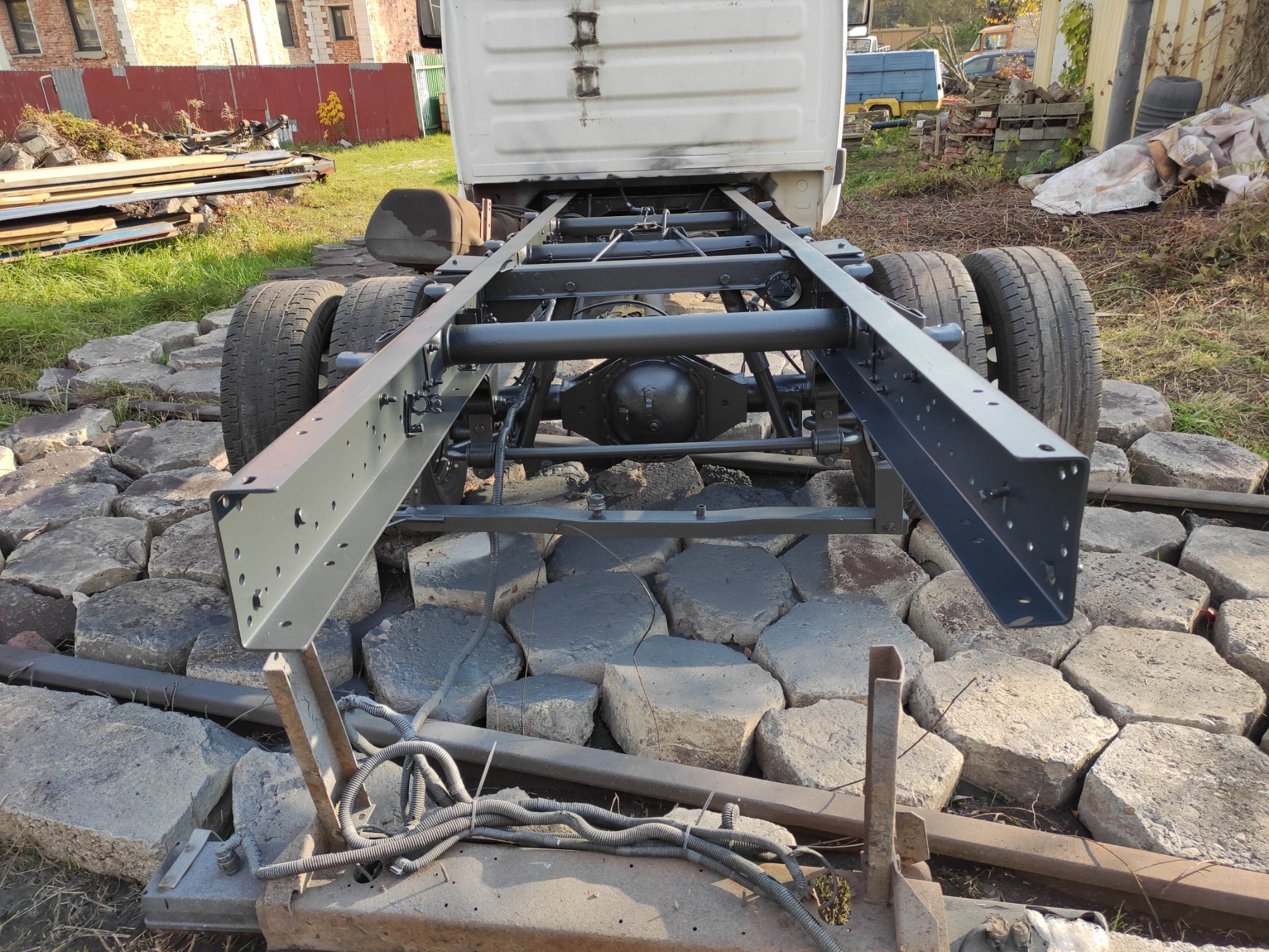 Surowa rama podwozia samochodu ciężarowego, pomalowana na szaro, stoi na nierównym podłożu z betonowych płyt. Widoczne opony, osie i elementy zawieszenia. W tle budynek i sterta złomu.