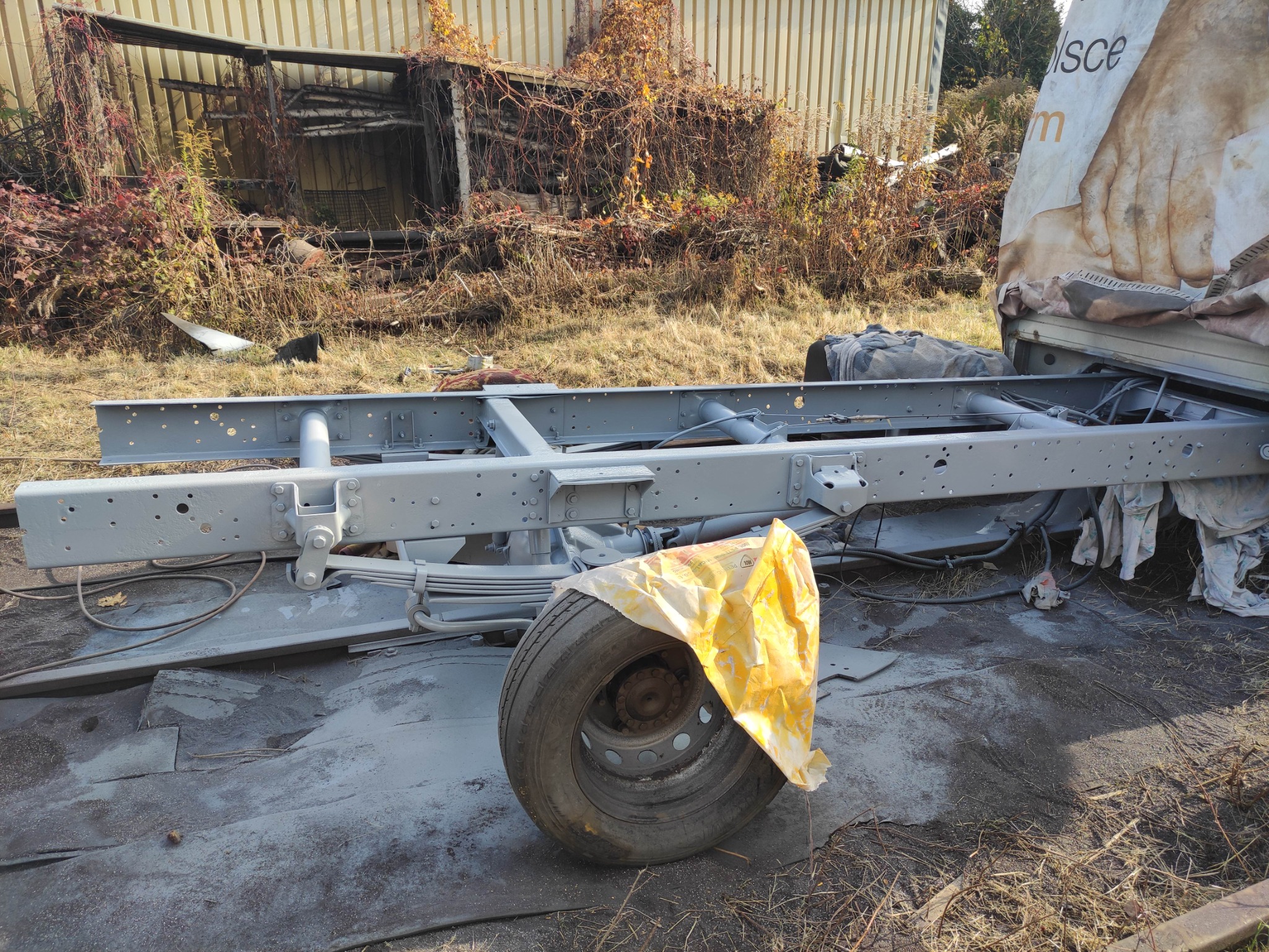 Szara, pomalowana rama samochodu ciężarowego z kołem zabezpieczonym folią, widoczna na zewnątrz w otoczeniu roślinności i elementów konstrukcyjnych.