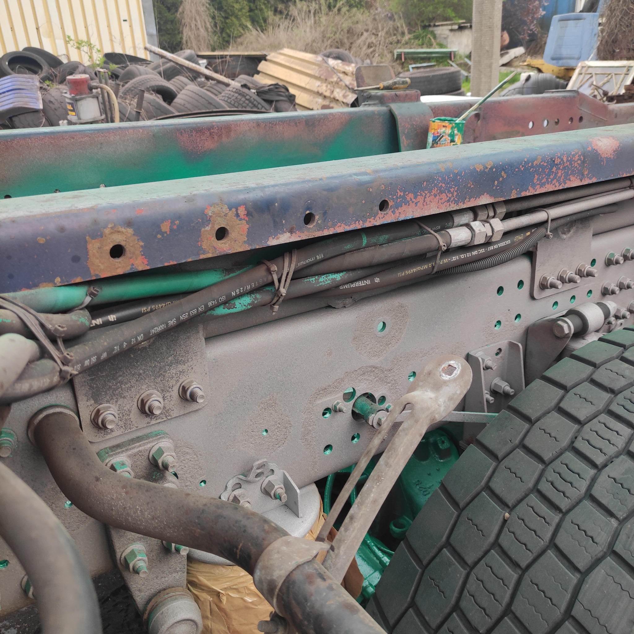 Fragment podwozia ciężarówki w trakcie naprawy, widoczne elementy metalowe, rury, opony i ślady malowania. W tle składowisko opon i inne elementy pojazdów.