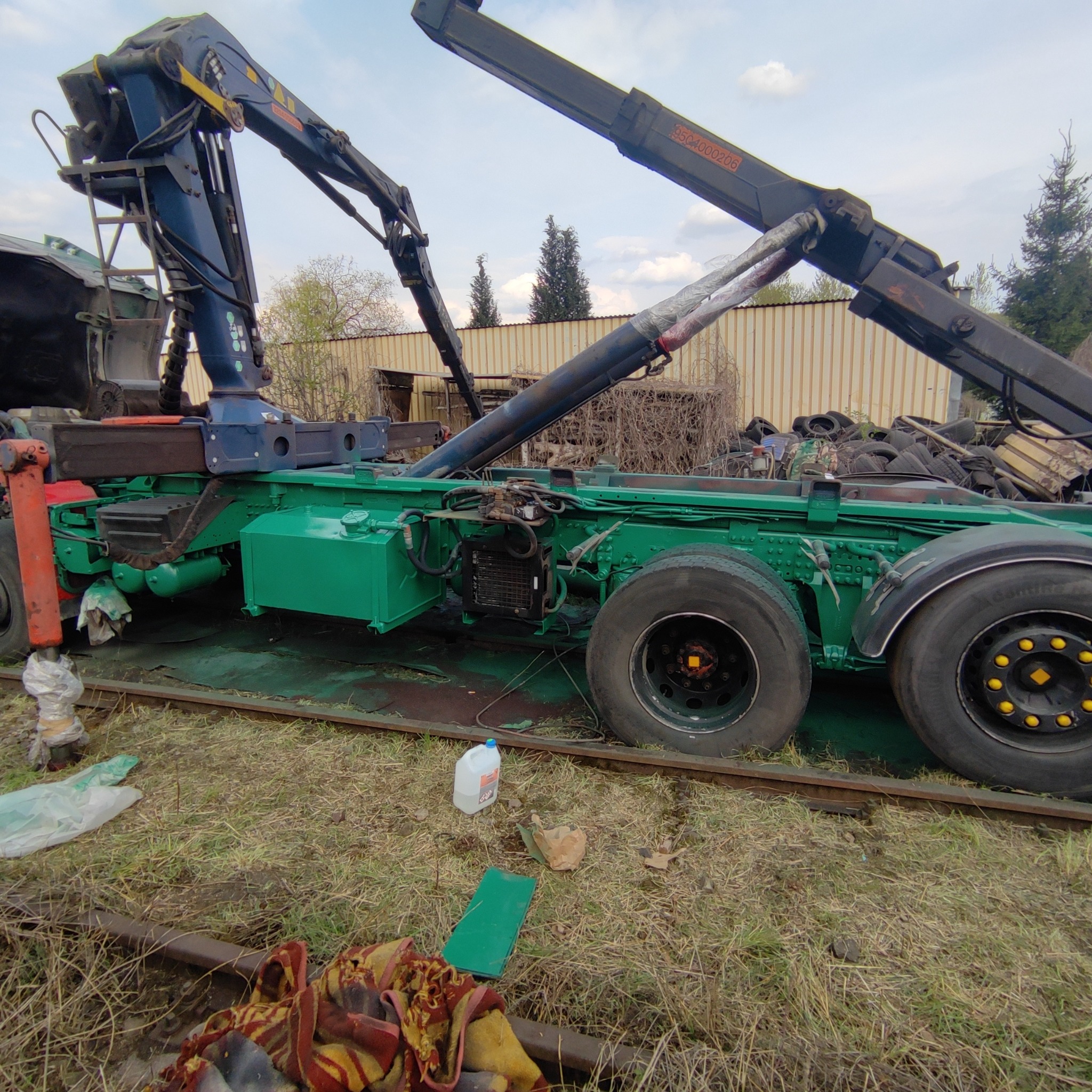 Zielona platforma ciężarówki z zamontowanymi ramionami dźwigowymi, stojąca na torach kolejowych, otoczona trawą i śmieciami, z widocznym zbiornikiem płynu i stertą opon w tle.