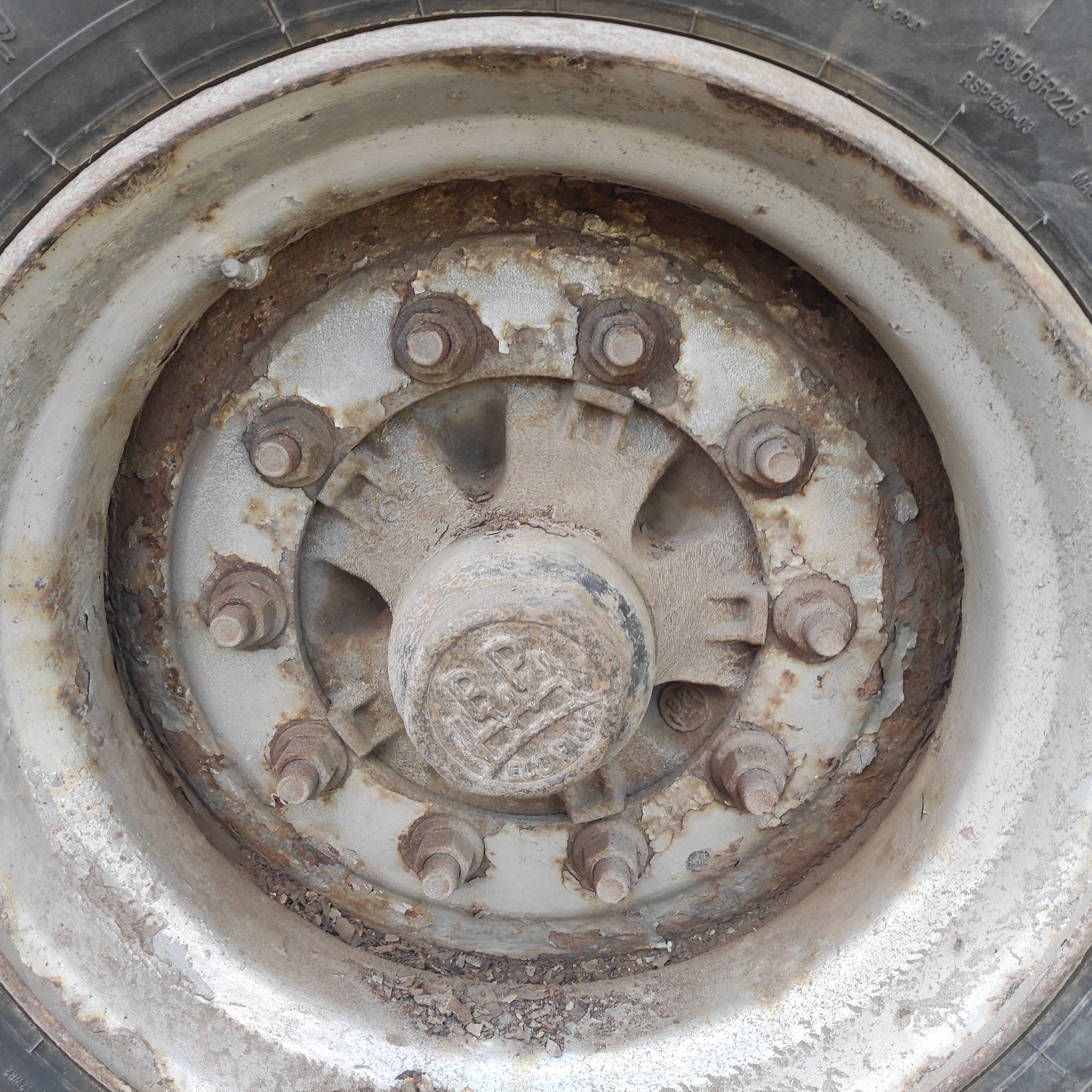 Zbliżenie na zardzewiałe, odrapane koło ciężarówki z widocznymi śrubami i oponą w tle, ukazujące zużycie i wpływ warunków atmosferycznych.