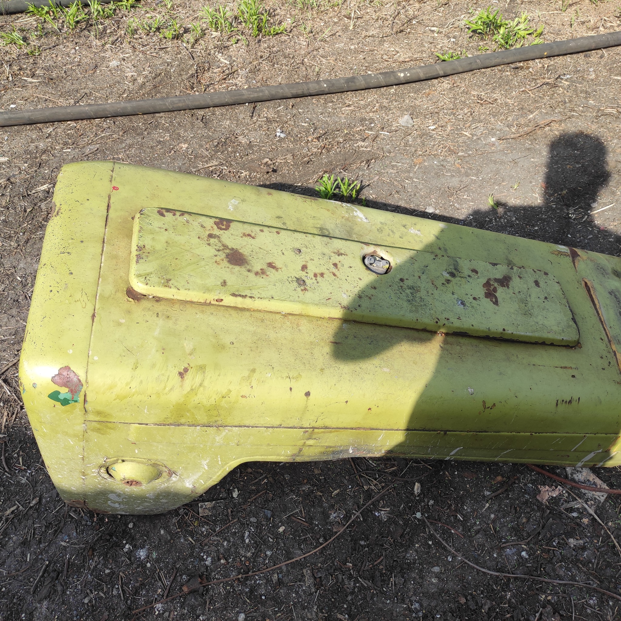 Zardzewiała, oliwkowo-zielona obudowa maszyny rolniczej z elementami mocującymi, leżąca na ziemi z fragmentami roślinności i czarnym wężem gumowym w tle.