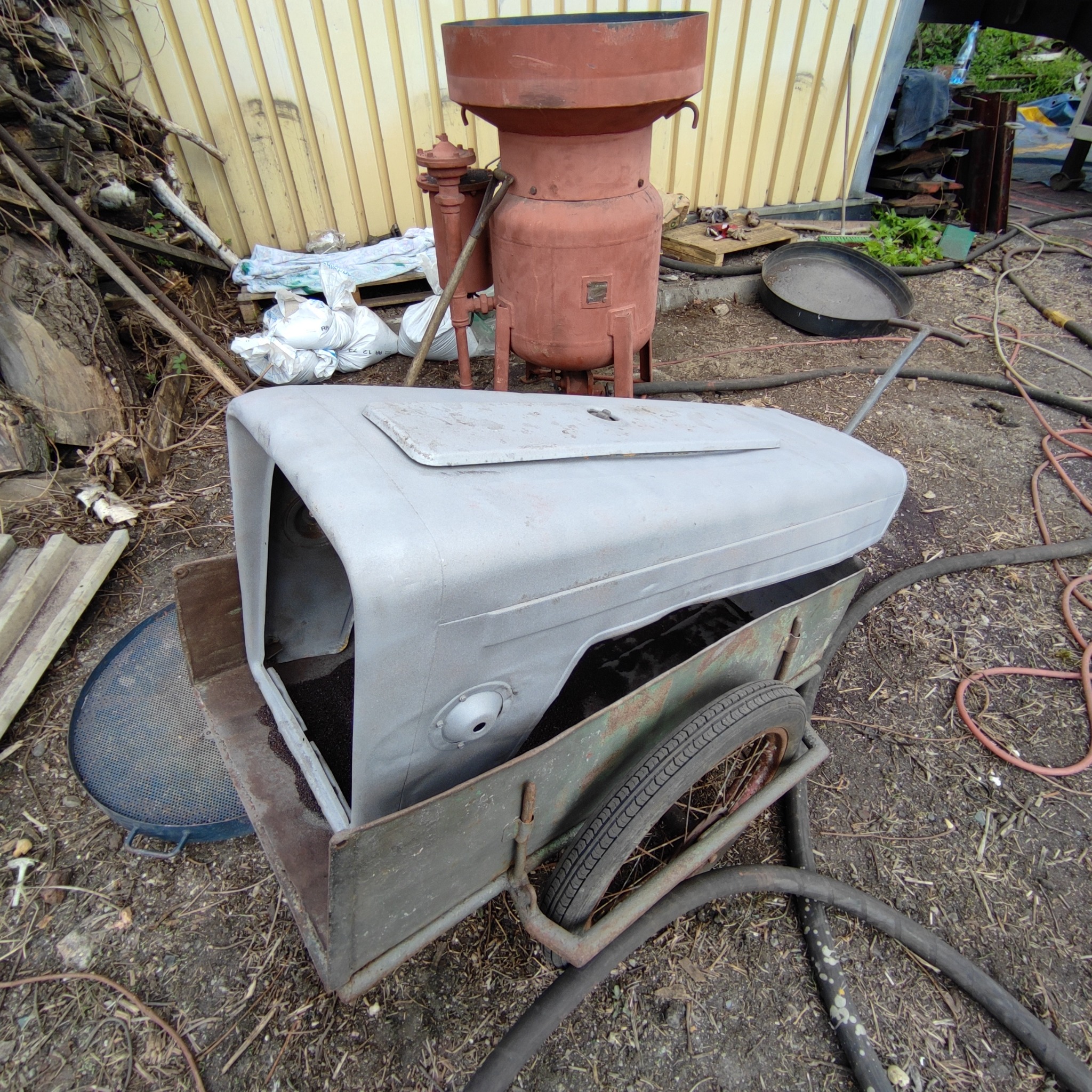 Szara, metalowa obudowa ciągnika rolniczego umieszczona na wózku transportowym z kołami, na ziemi widoczne węże i elementy metalowe, w tle zbiornik ciśnieniowy.