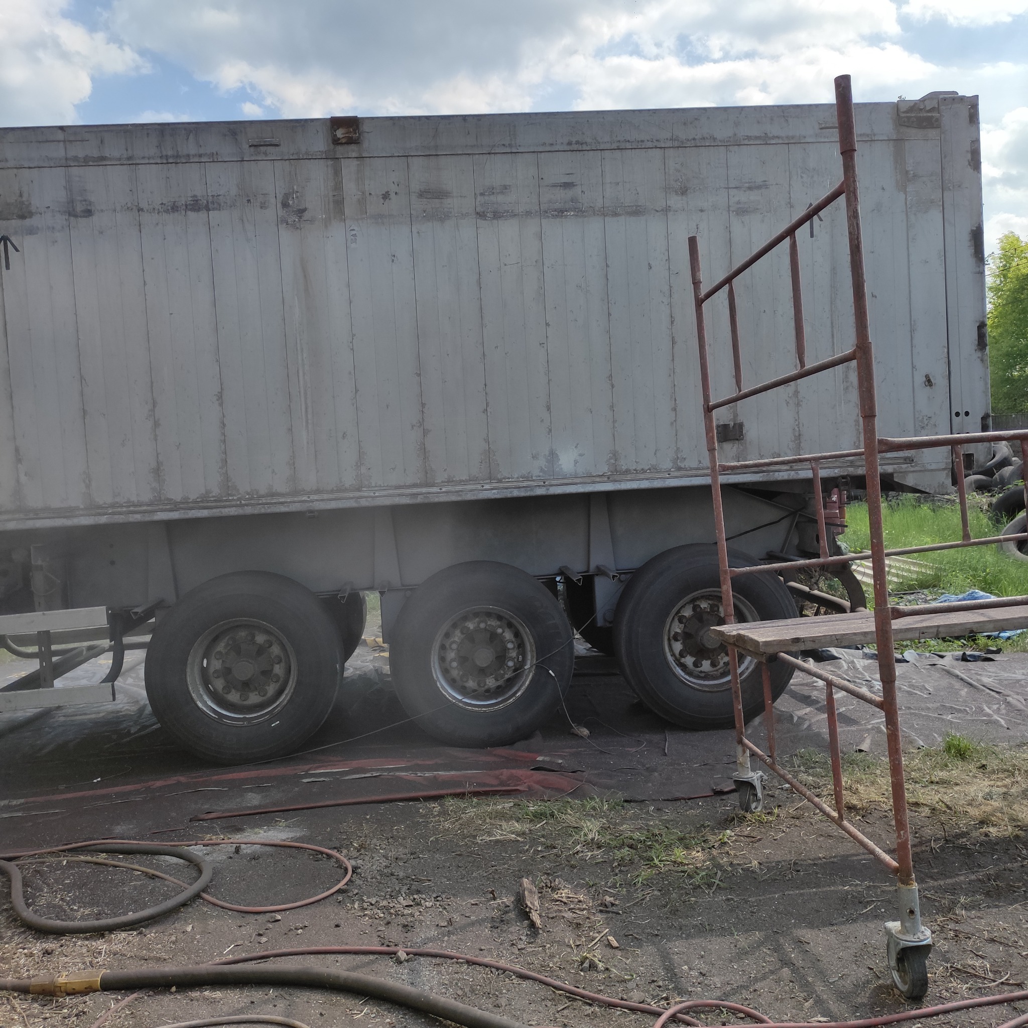 Przyczepa ciężarowa częściowo osłonięta rusztowaniem, prawdopodobnie w trakcie czyszczenia lub przygotowywania do malowania, z widocznymi śladami rdzy i zabrudzeń, oraz leżącymi na ziemi przewodami.