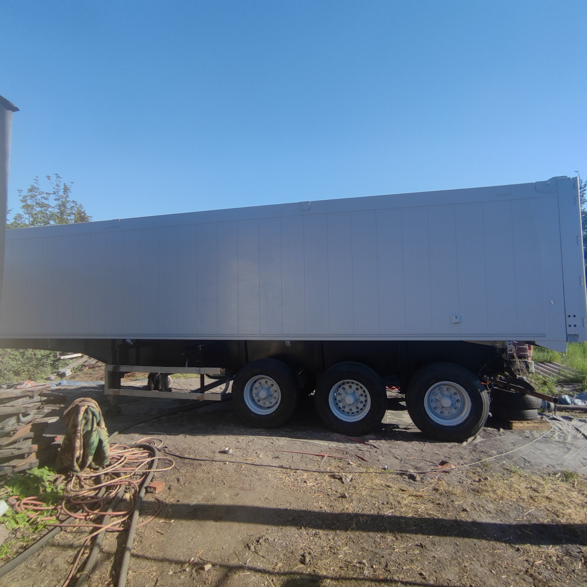 Długa, szara naczepa samochodowa z trzema osiami kół, stojąca na ziemi z widocznymi przewodami i elementami metalowymi w tle, sfotografowana w słoneczny dzień.