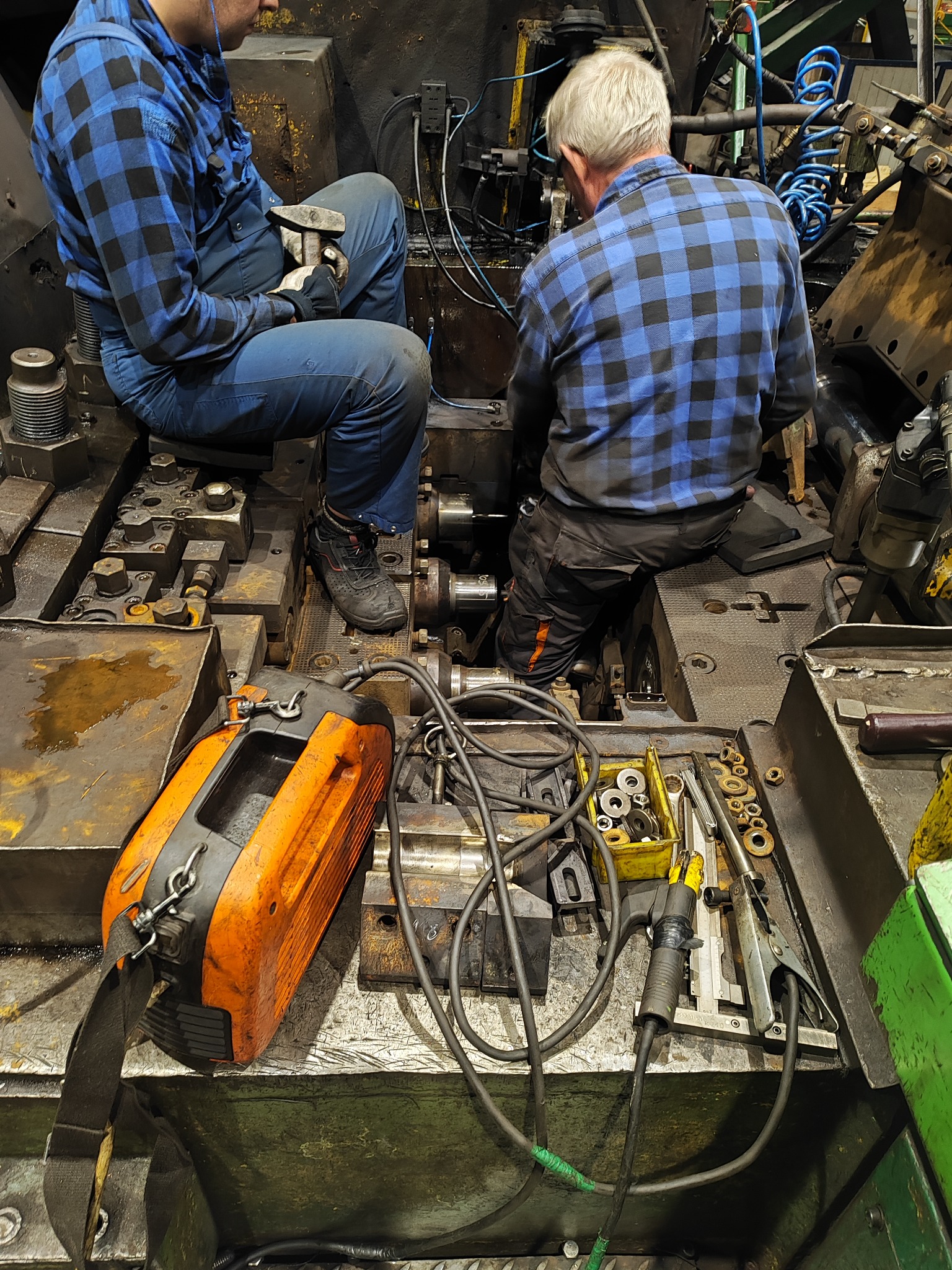 Dwóch mężczyzn w niebieskich koszulach w kratę siedzi na dużej, metalowej konstrukcji, prawdopodobnie maszynie przemysłowej, podczas naprawy lub konserwacji. W tle widoczne przewody i elementy...