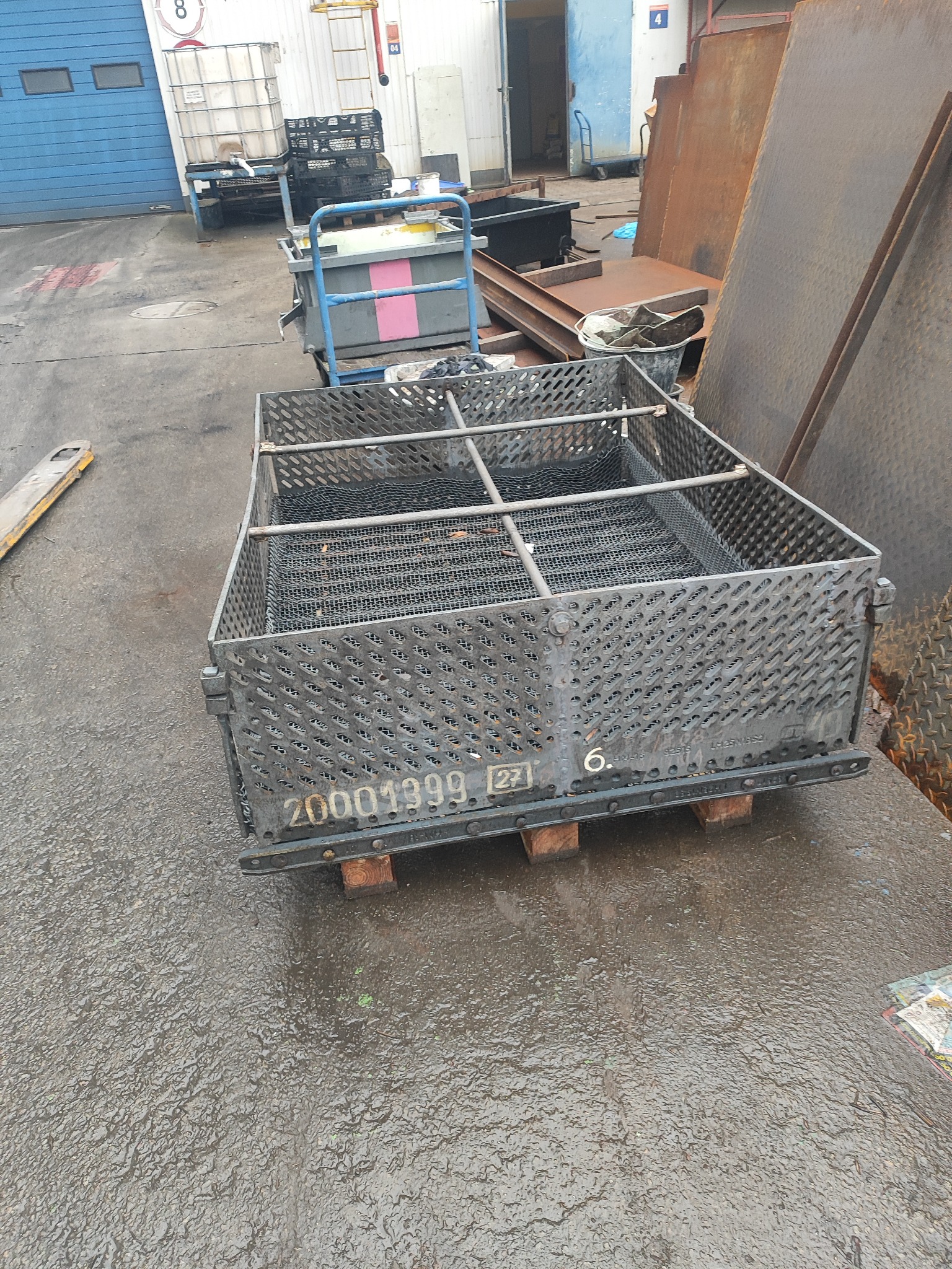 Metalowa ażurowa skrzynia z numerem seryjnym 20001399 i oznaczeniem 6.27, stojąca na drewnianych paletach na mokrym betonowym podłożu, w tle zabudowania przemysłowe i inne konstrukcje metalowe.