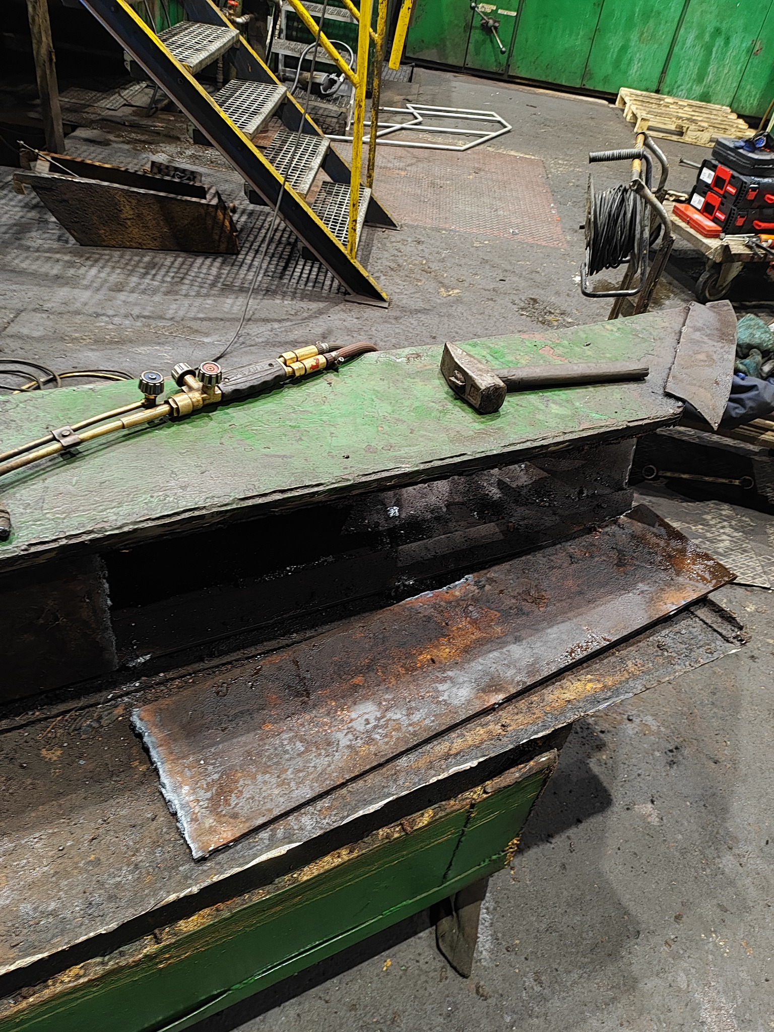 Palnik acetylenowo-tlenowy, młotek i przycięty arkusz metalu na zielonym stole warsztatowym w hali produkcyjnej, w tle schody i wózek z narzędziami.