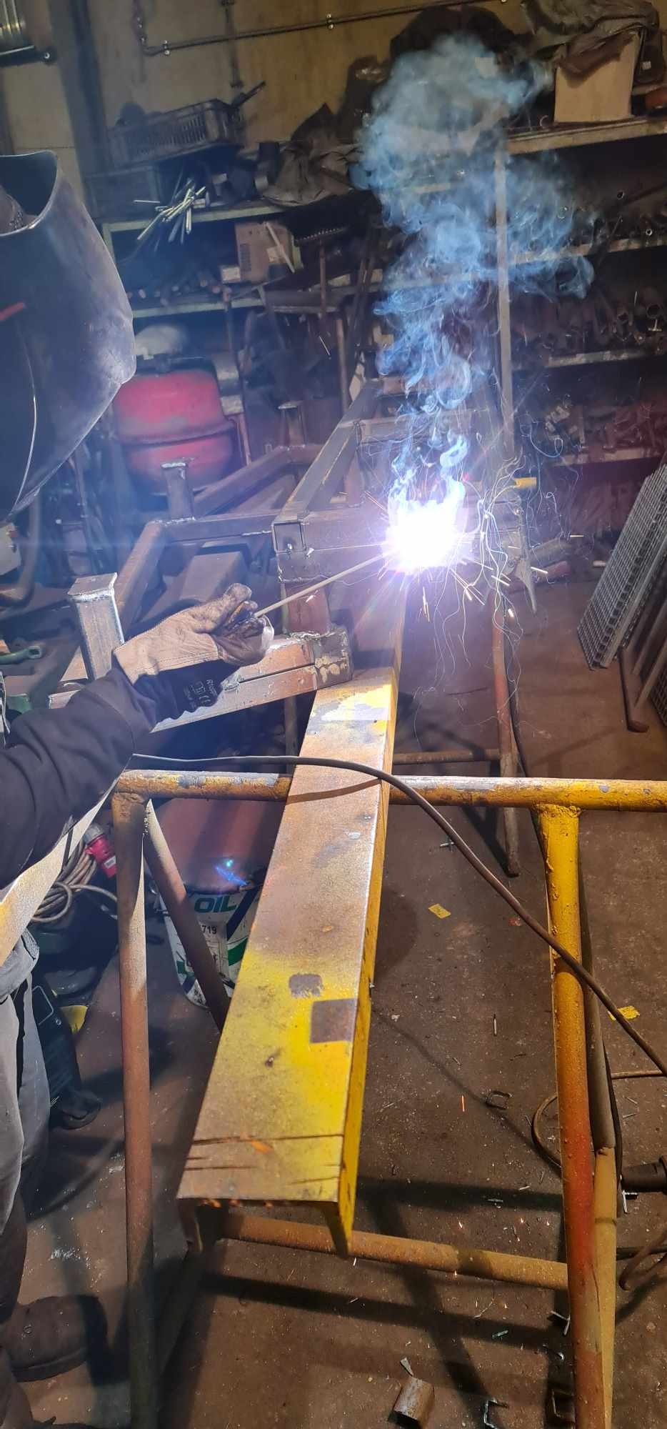 Prace montażowe konstrukcji stalowej: spawanie metalowych elementów ramy w warsztacie, widoczne iskry i dym, pracownik w masce i rękawicach ochronnych.