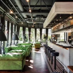 Wnętrze baru z zielonymi sofami Chesterfield, wysokimi stołkami barowymi i oknami od podłogi do sufitu, oświetlone industrialnymi lampami wiszącymi.