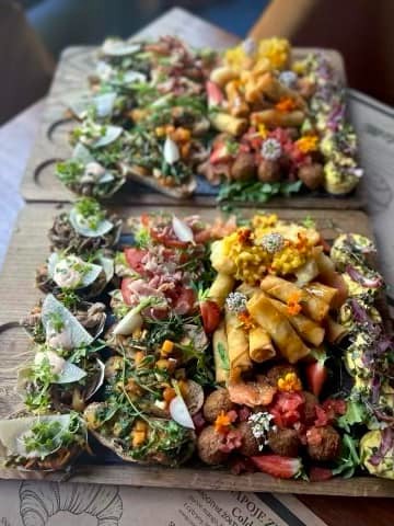 Dwa drewniane talerze z różnorodnymi przekąskami: mini kanapeczki z warzywami i serem, roladki, kulki mięsne, dekorowane jadalnymi kwiatami i ziołami.