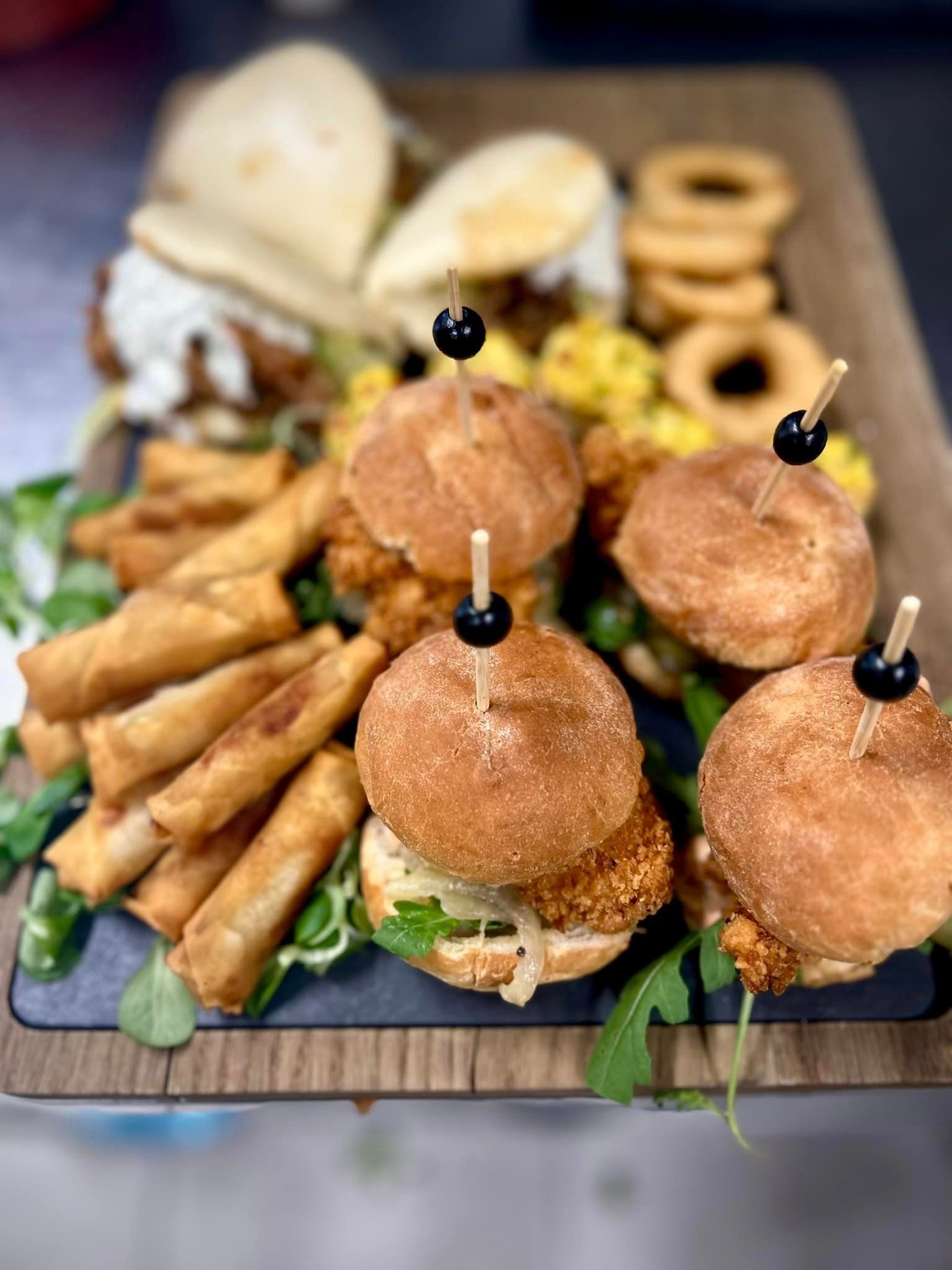 Drewniana taca z różnorodnymi przekąskami: mini burgery z kotletem w panierce i czarną oliwką na wykałaczce, sajgonki, krążki cebulowe i mini bułeczki z sosem, całość udekorowana świeżą rukolą...