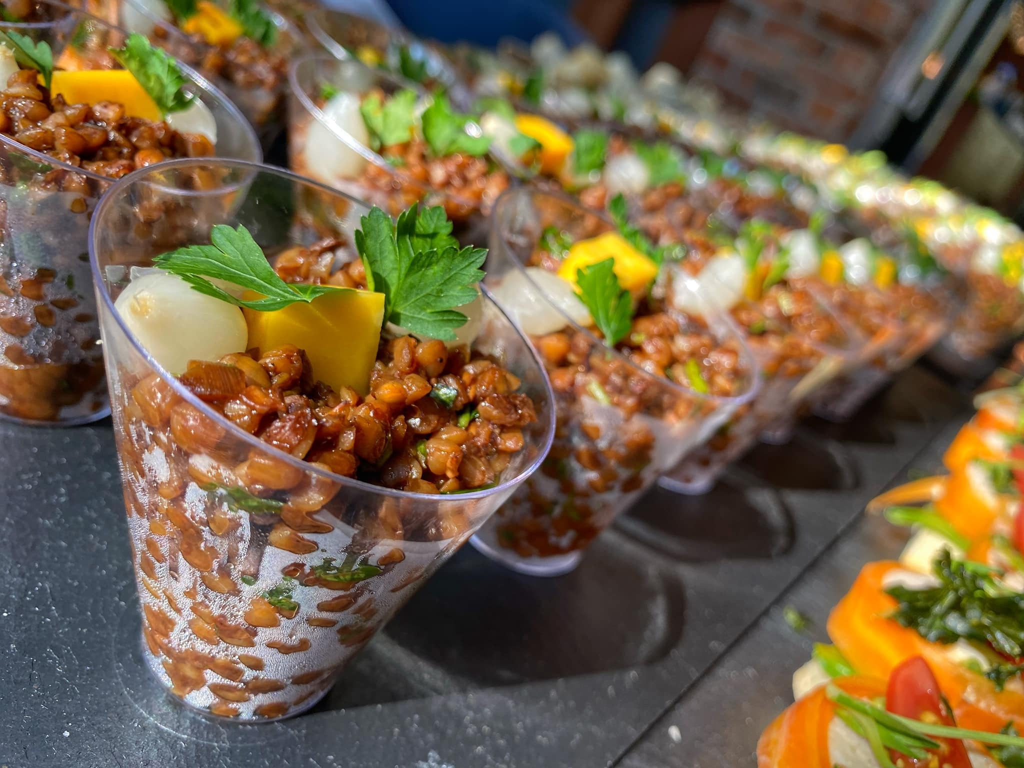 Ustawione w rzędzie porcje brązowej sałatki w plastikowych kubeczkach, udekorowane zieloną pietruszką, białymi cebulkami i kostką żółtego sera, na metalowej powierzchni.