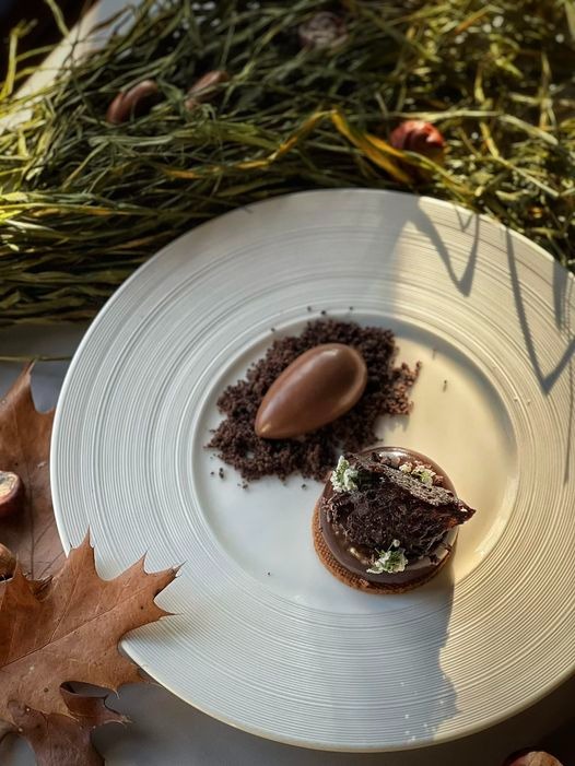 Elegancko podany deser na białym talerzu z dekoracją z jesiennych liści i gałązek, czekoladowe ciastko z kremem i polewą, obok czekoladowa łza na kruszonce.