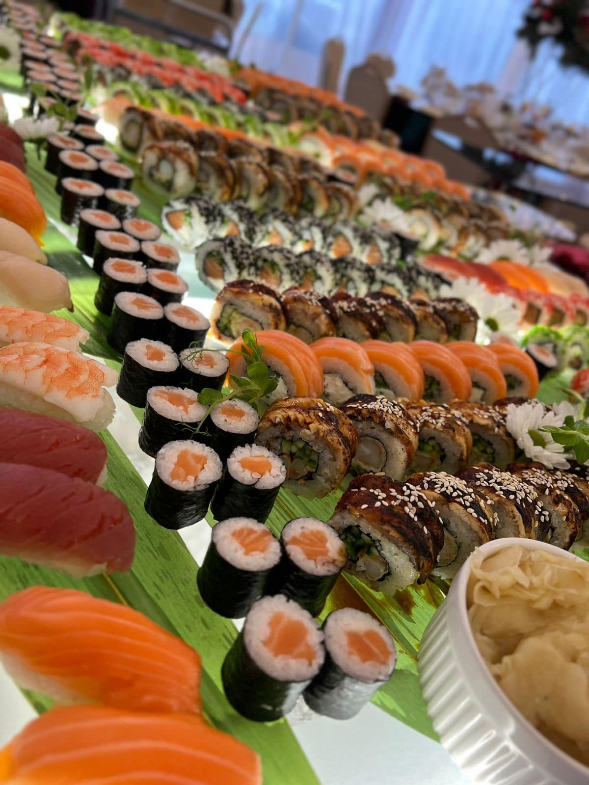 Bogaty wybór sushi, w tym maki z łososiem, nigiri z tuńczykiem i krewetkami, oraz rolki z węgorzem i sezamem, ułożone na zielonych liściach i serwowane z imbirem.
