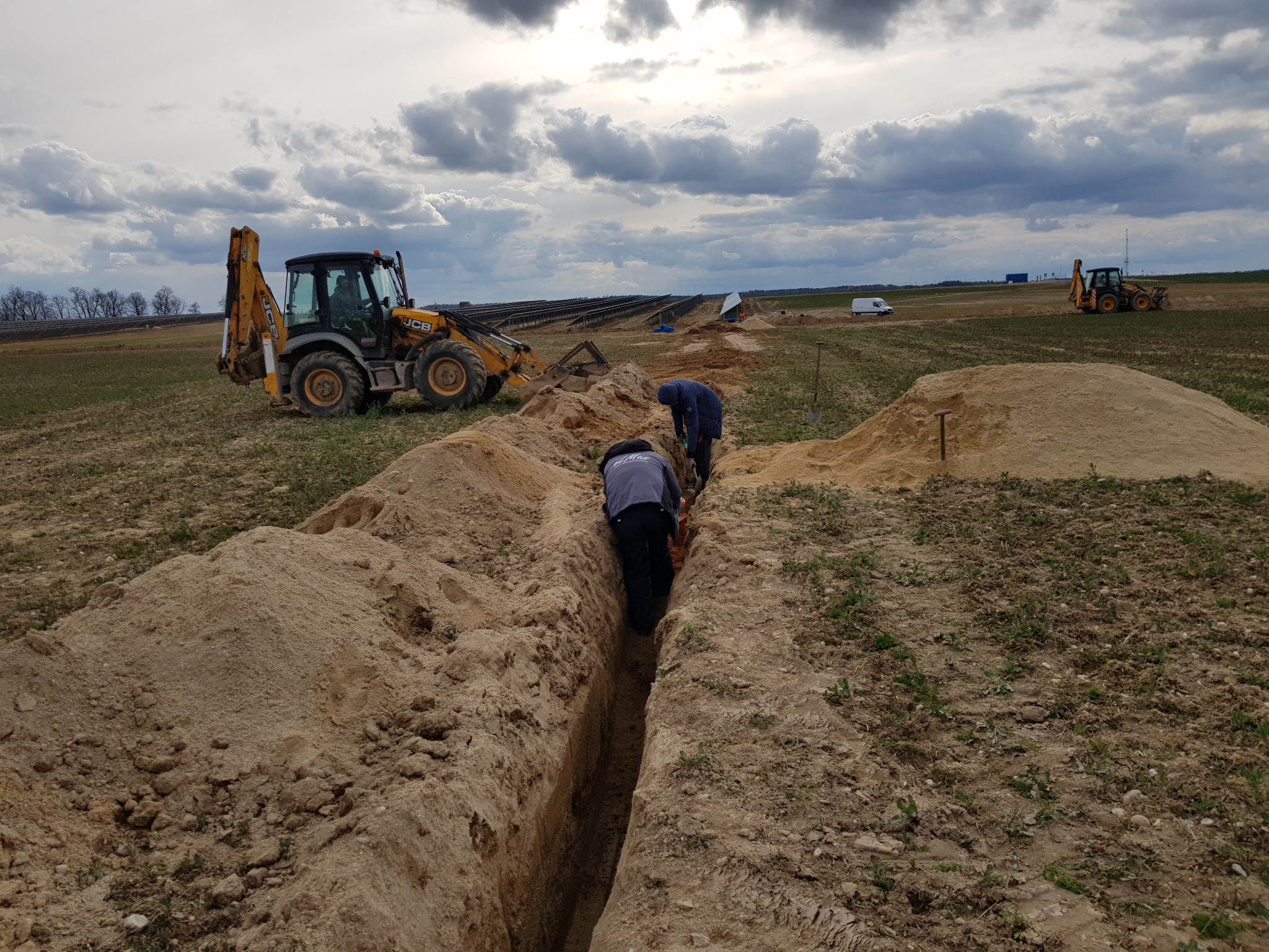 Dwaj robotnicy kopią wąski rów w ziemi na farmie fotowoltaicznej, w tle widoczny żółty traktor JCB oraz panele słoneczne i pochmurne niebo.