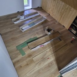 Krzysztof Bączyński - Podczas układania paneli podłogowych na poddaszu, widoczne narzędzia, paczki paneli i fragment buta osoby wykonującej pracę. Ukończony fragment podłogi.