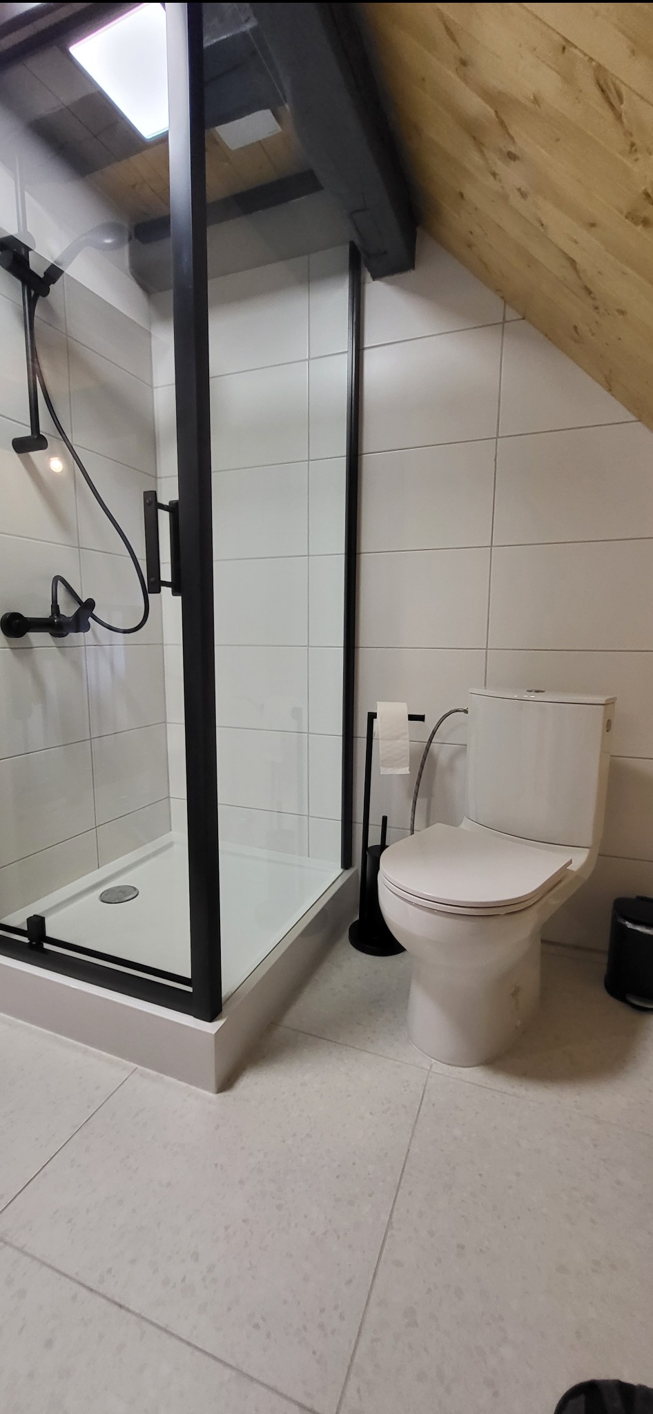 Nowoczesna łazienka na poddaszu z kabiną prysznicową w czarnej ramie, białą toaletą i jasnymi płytkami. Uwagę przykuwa drewniana konstrukcja dachu.