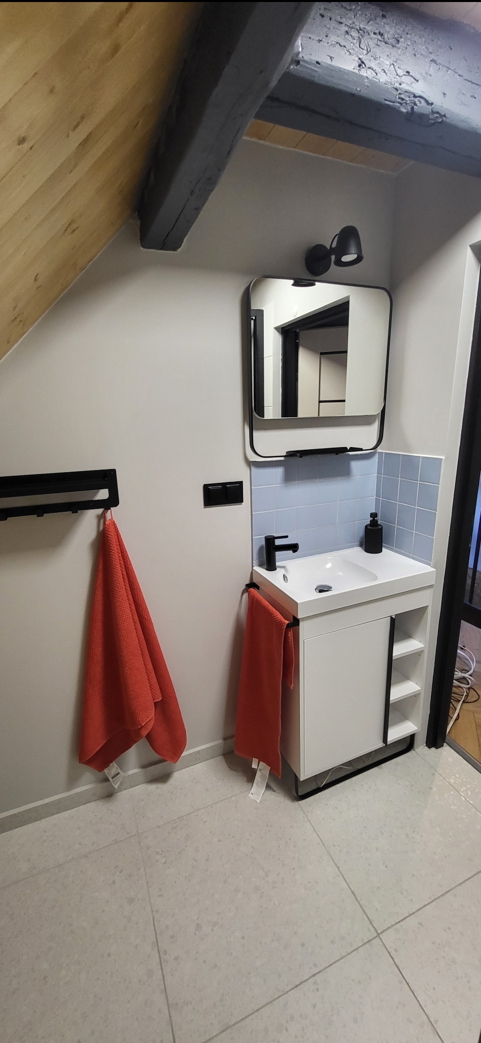 Wykończona łazienka na poddaszu: biała szafka z umywalką i czarną armaturą, lustro w czarnej ramie, niebieskie płytki, pomarańczowe ręczniki i drewniane belki stropowe.