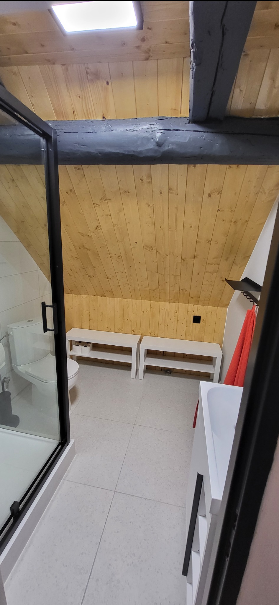 Nowoczesna łazienka na poddaszu z drewnianą boazerią, kabiną prysznicową z czarną ramą i białymi meblami. Ukośny sufit z belkami.