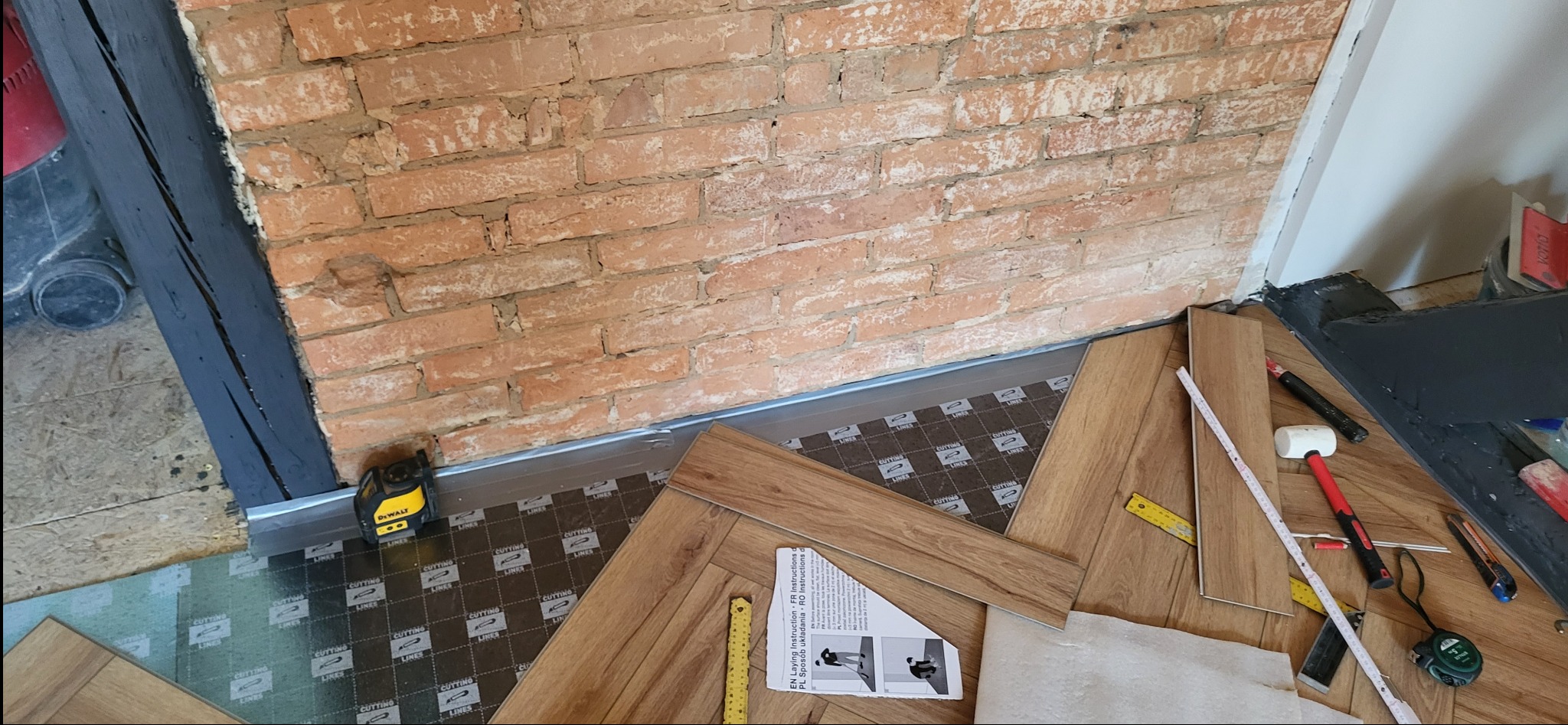 Układanie paneli podłogowych w pomieszczeniu z ceglaną ścianą. Widoczne narzędzia, listwa przypodłogowa i instrukcja montażu. Laserowy niwelator DeWalt ustawiony przy ścianie.
