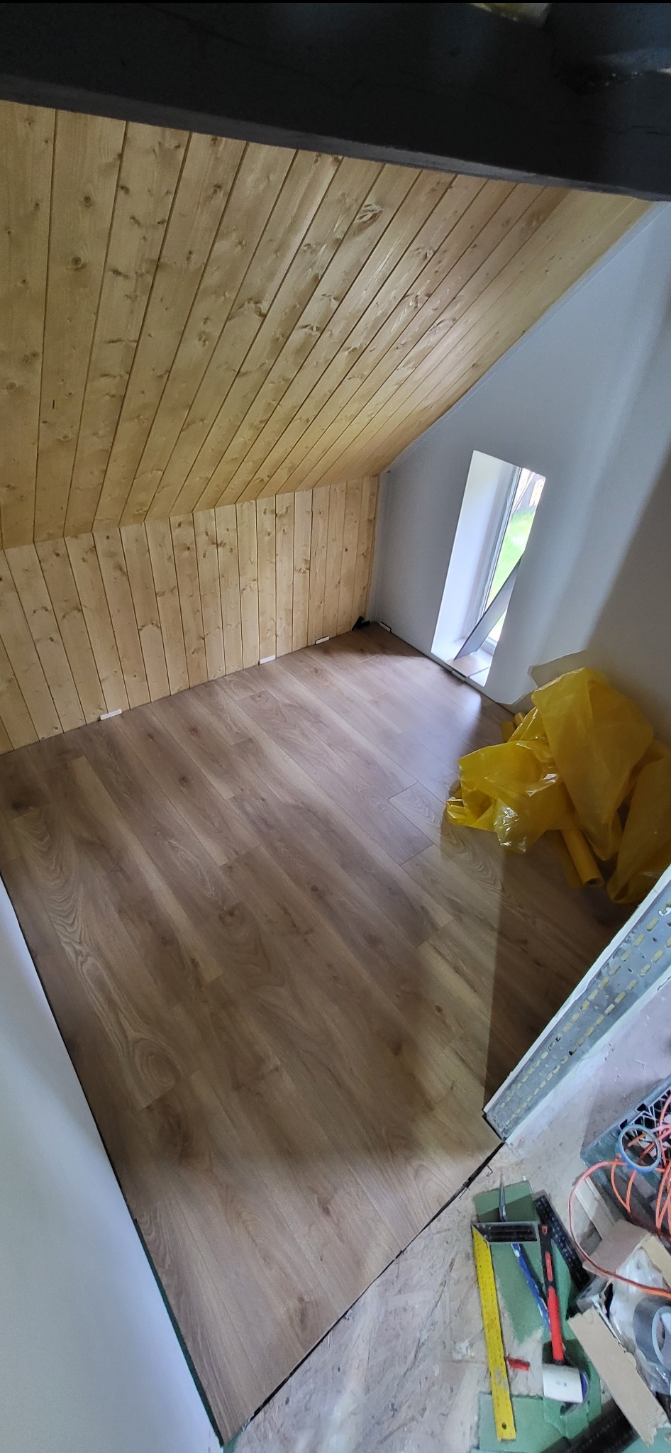 Pokój na poddaszu z drewnianą boazerią i panelami podłogowymi w trakcie montażu. Widoczne narzędzia i materiały budowlane.