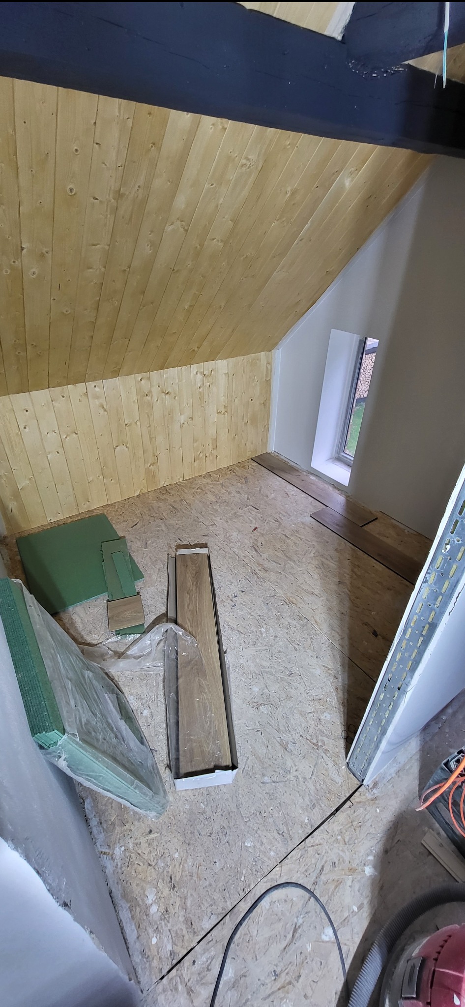 Poddasze w trakcie wykańczania: drewniana boazeria, płyty OSB na podłodze, panele w opakowaniu, mata wygłuszająca. Fragment okna i narzędzia w kadrze.
