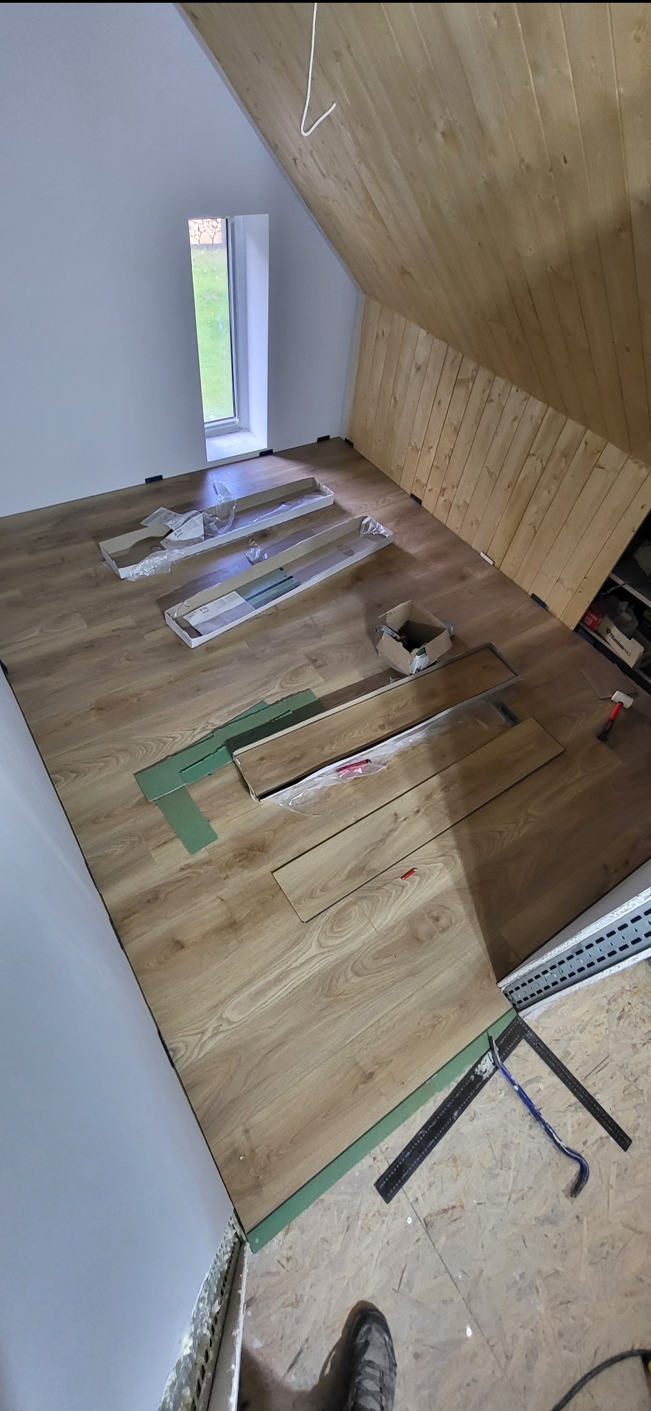 Podczas układania paneli podłogowych na poddaszu, widoczne narzędzia, paczki paneli i fragment buta osoby wykonującej pracę. Ukończony fragment podłogi.