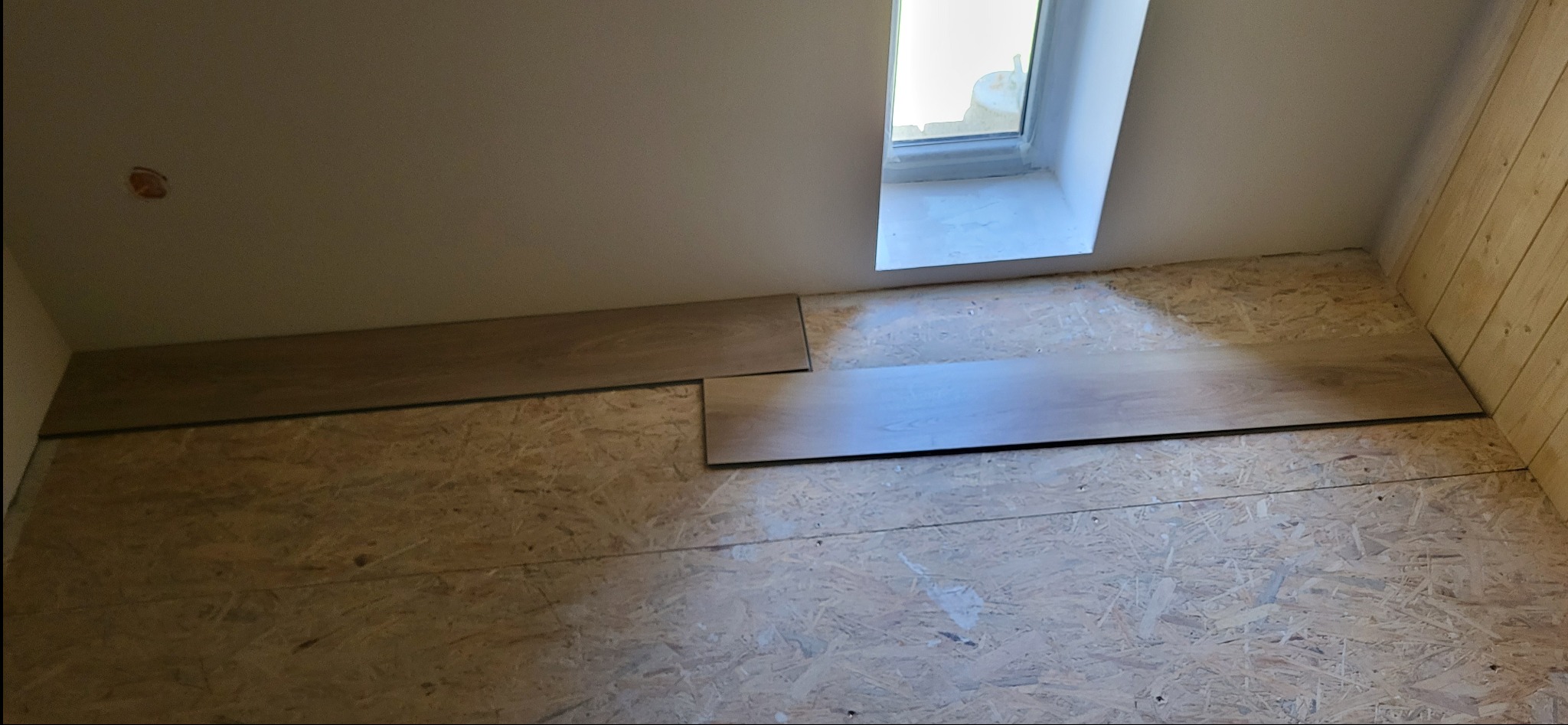 Fragment podłogi z płyt OSB, na której układane są panele podłogowe w jasnym odcieniu drewna. Widoczne okno i drewniana boazeria na ścianie.