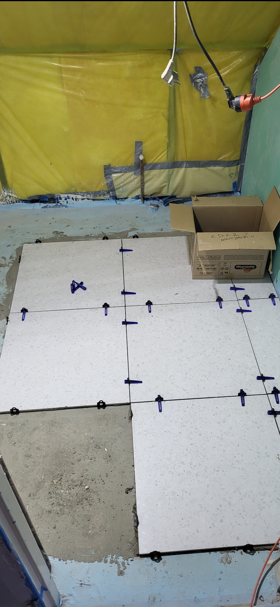 Układanie jasnych płytek podłogowych z użyciem systemu poziomowania; widoczna kartonowa skrzynka i folia ochronna na ścianach. Fragment podłogi bez płytek.