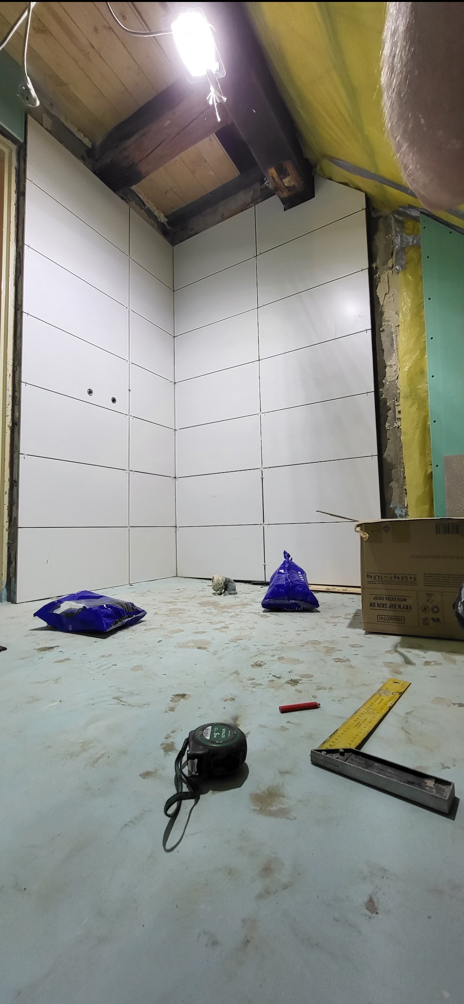 Białe płytki ułożone na ścianach pod skosem w trakcie remontu łazienki, widoczne narzędzia i worki z materiałami budowlanymi na podłodze.