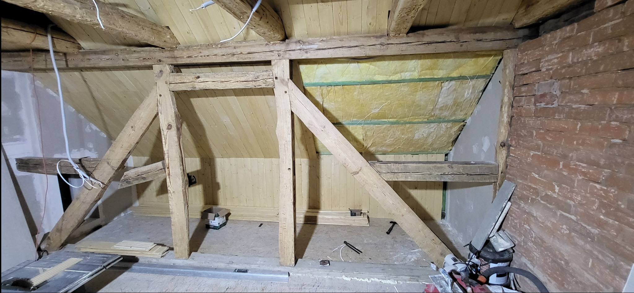Adaptacja poddasza: drewniana konstrukcja, ocieplenie wełną mineralną, boazeria na ścianach, fragment ściany z cegły. Widoczne narzędzia i materiały budowlane.