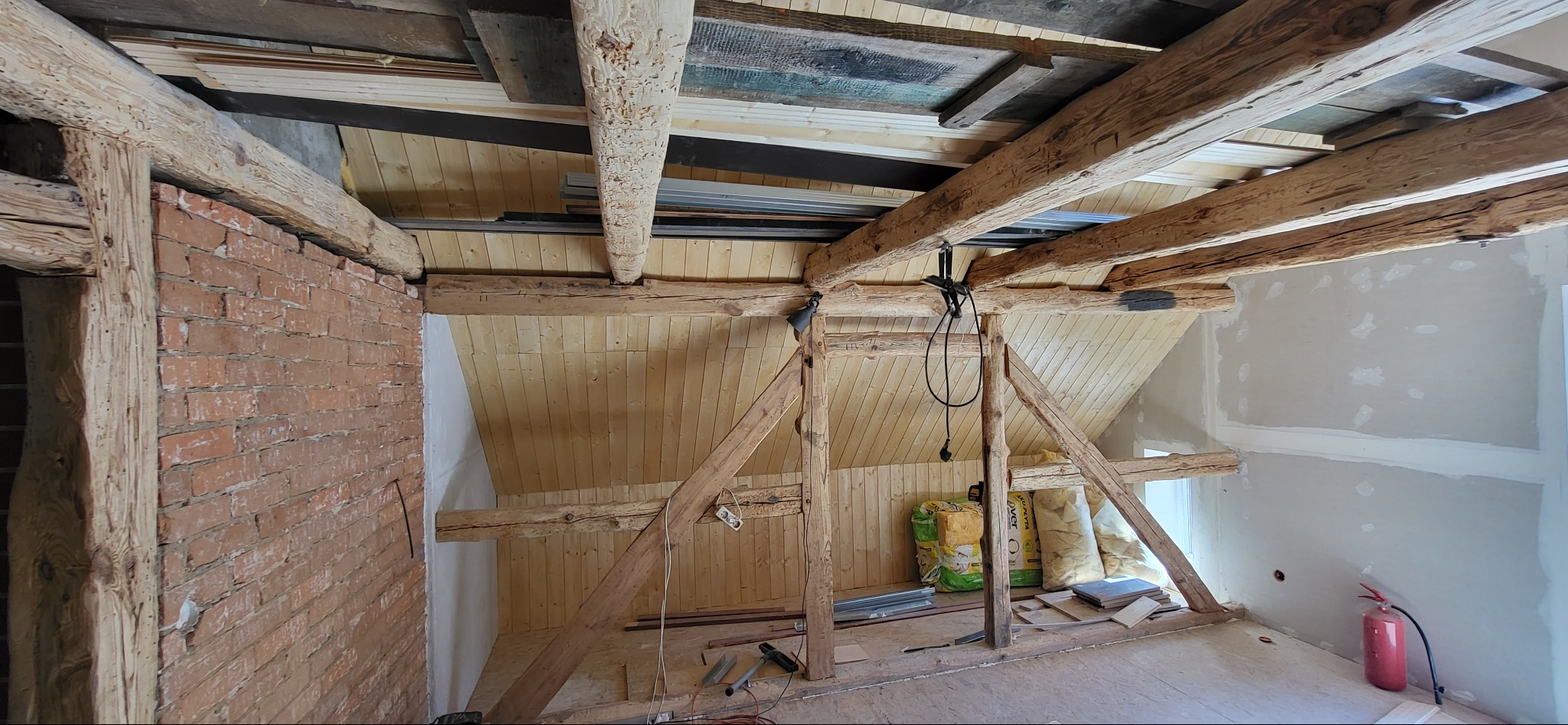 Poddasze w trakcie adaptacji: drewniane belki konstrukcyjne, fragment ściany z cegły, płyty OSB na podłodze, ocieplenie w workach. Widoczne narzędzia i gaśnica.