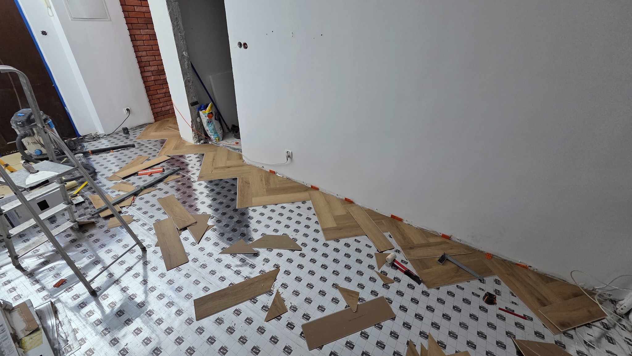 Układanie paneli w jodełkę na podkładzie z folii. Widoczne narzędzia, drabina i odkurzacz. Częściowo ułożona podłoga w jasnym odcieniu drewna.
