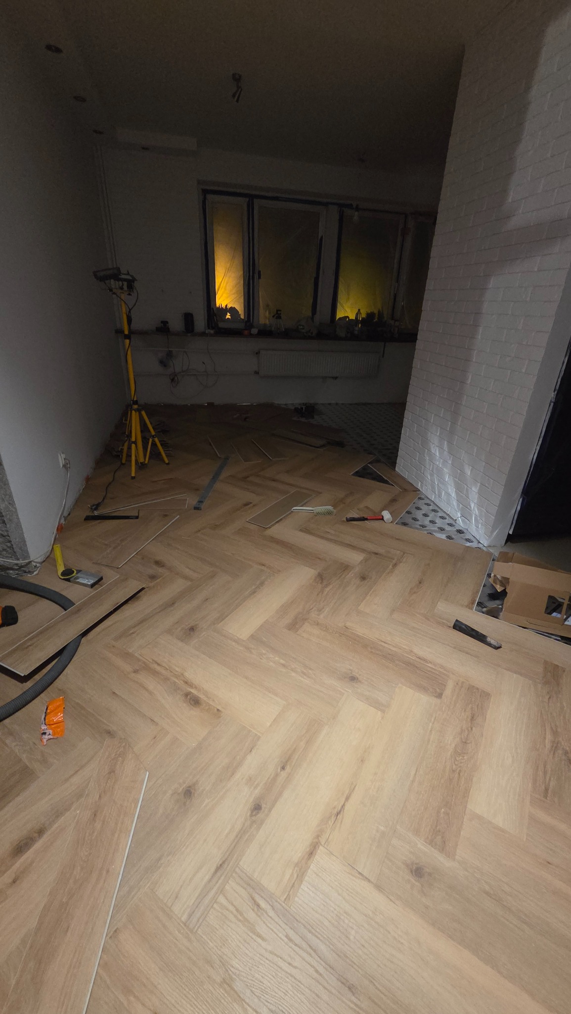 Układanie paneli podłogowych w jodełkę, widoczne narzędzia i lampa oświetleniowa. Częściowo wykończona podłoga w pomieszczeniu z oknem i ścianą z cegły.