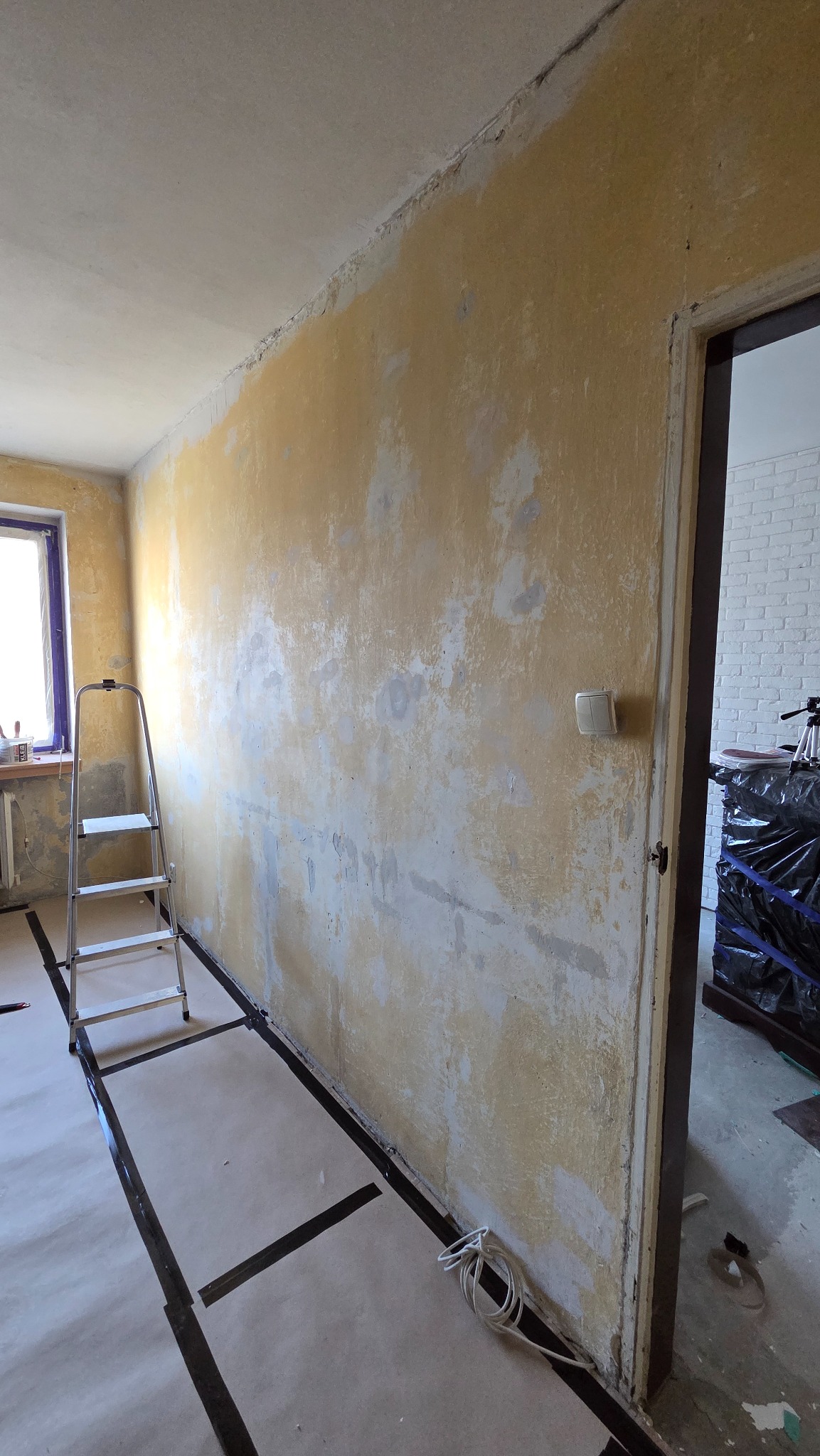 Pokój w trakcie przygotowań do malowania: zabezpieczona podłoga, drabina oparta o ścianę z widocznymi śladami szpachlowania. Widoczne okno i otwarte drzwi.