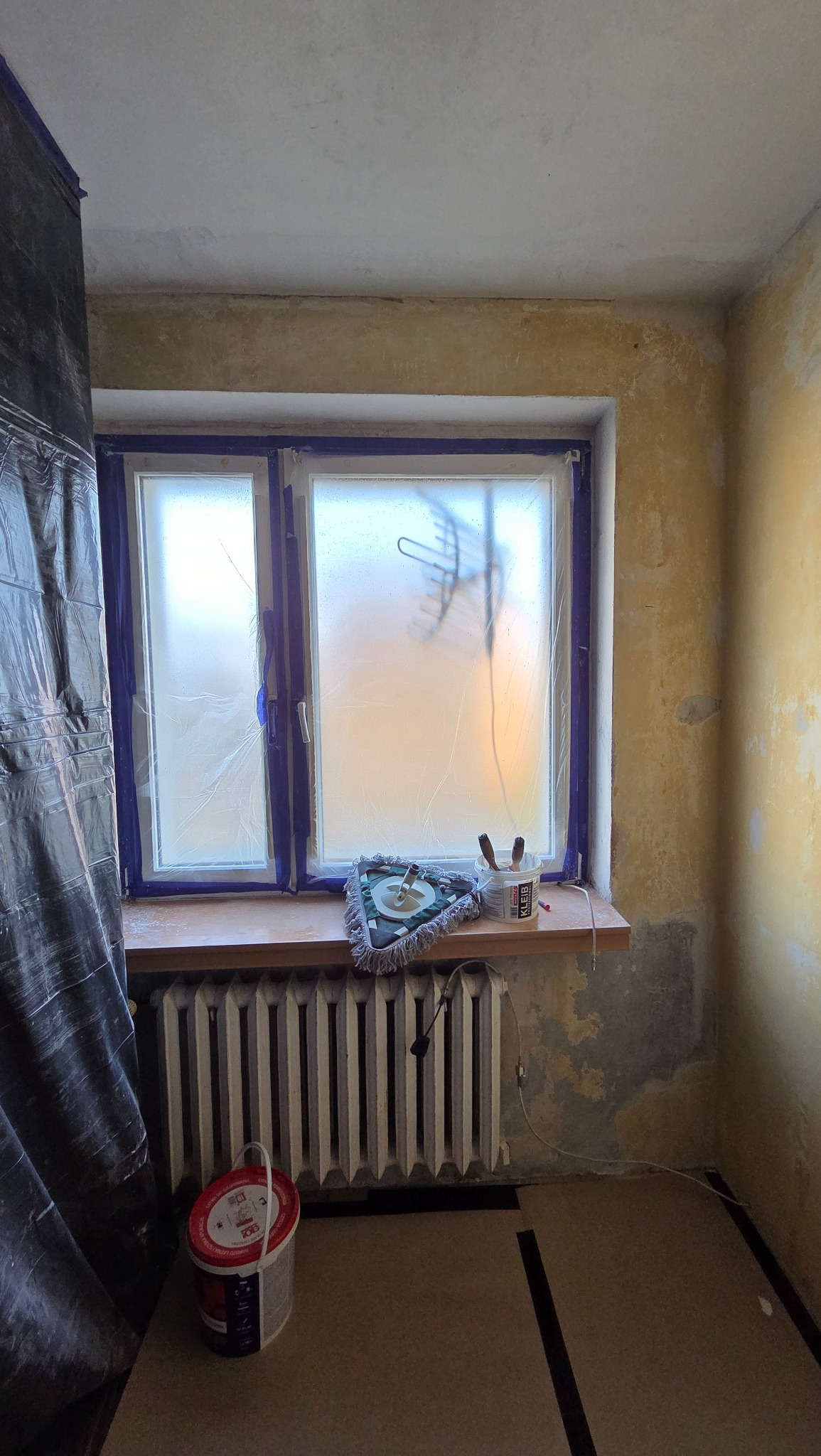 Pokój w trakcie przygotowań do malowania: okno zabezpieczone folią, ściany z widocznymi śladami starej farby, grzejnik, wiadro z farbą i narzędzia malarskie.