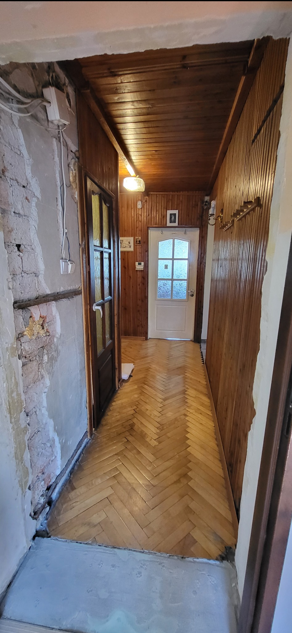 Korytarz w trakcie remontu: drewniana boazeria, parkiet w jodełkę, surowa ściana z odsłoniętą cegłą. Widoczne przewody elektryczne i przygotowania do dalszych prac wykończeniowych.