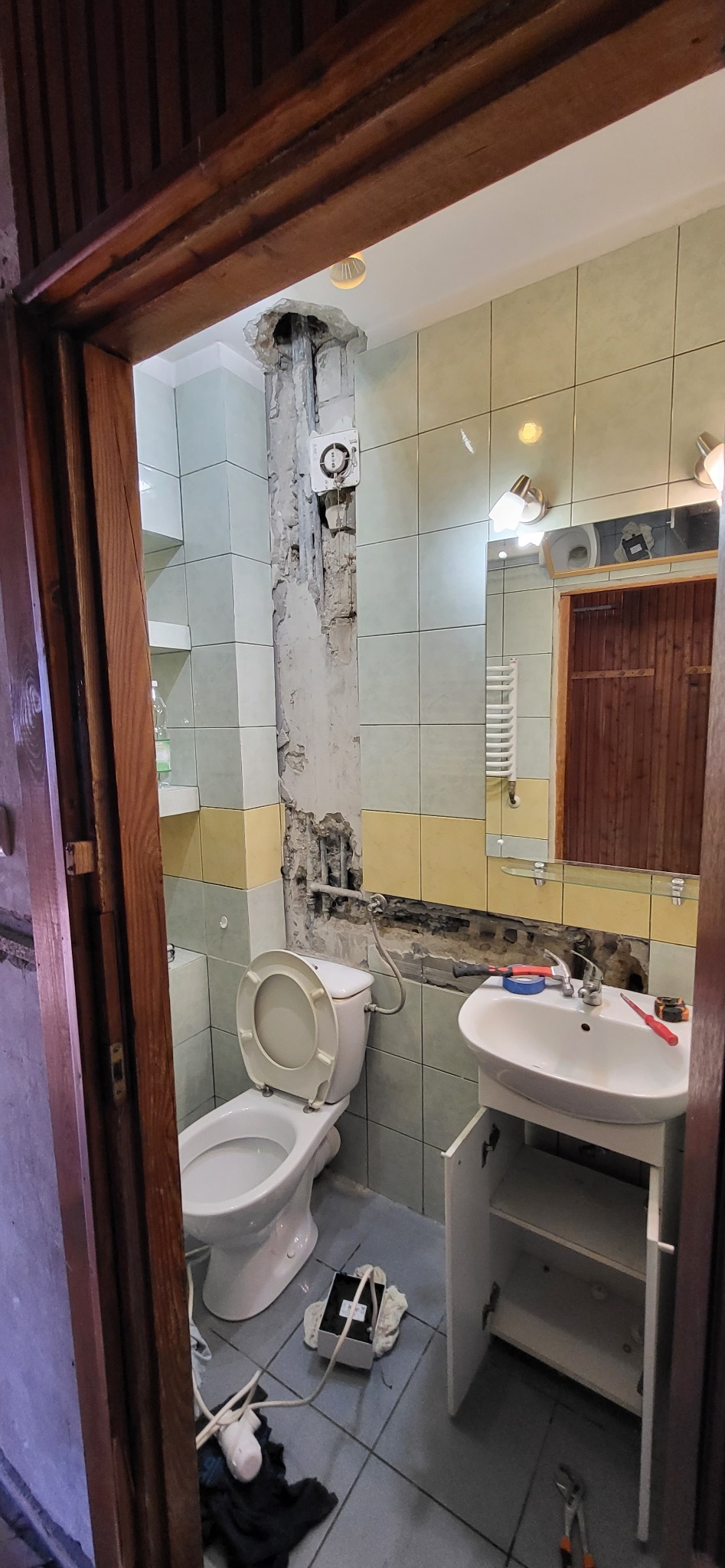 Łazienka w trakcie remontu: usunięte płytki, odsłonięta instalacja, widoczne narzędzia, toaleta i umywalka z szafką. Widok zza drewnianej futryny.