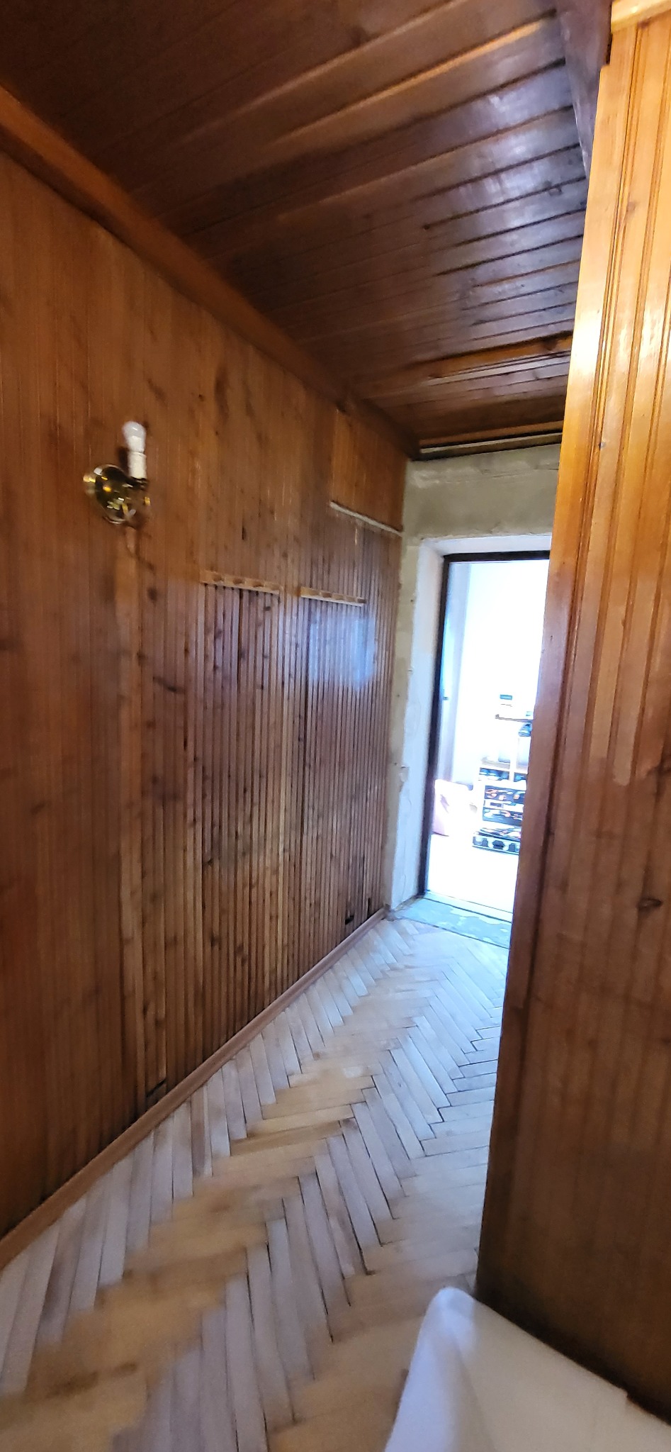 Korytarz wykończony boazerią w ciepłych odcieniach brązu, z parkietem ułożonym w jodełkę. Widoczna lampa ścienna i otwarte drzwi do innego pomieszczenia.