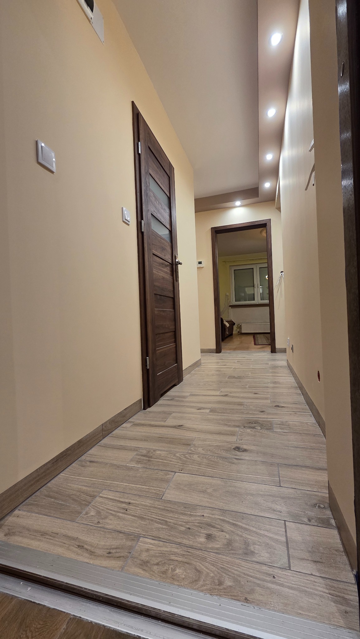 Korytarz po remoncie z drewnopodobną podłogą z płytek, beżowe ściany i ciemne drzwi z przeszkleniami. Sufit z oświetleniem punktowym. Widok na pokój.