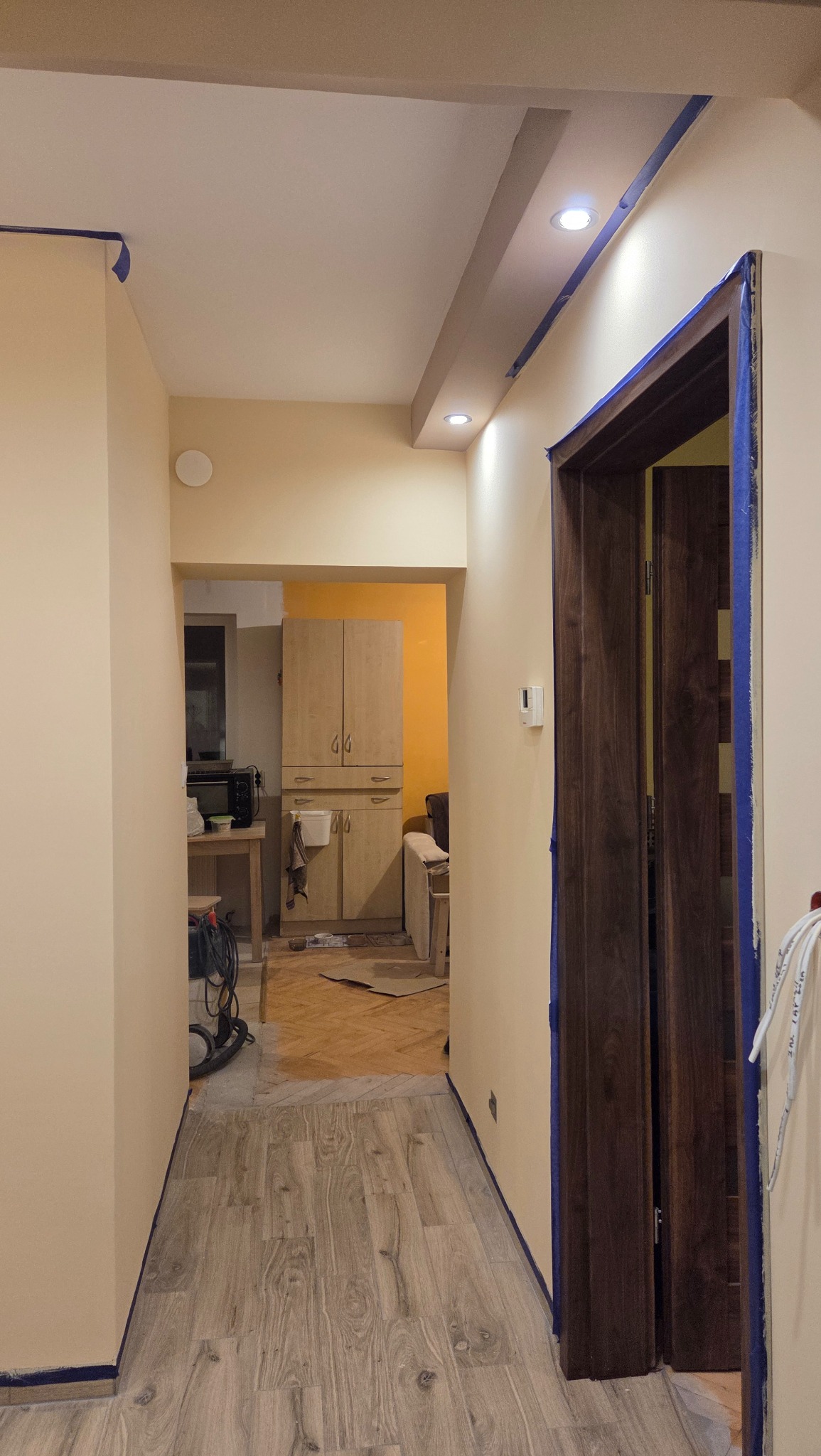 Korytarz w trakcie malowania, ściany zabezpieczone taśmą malarską, widoczne nowe panele podłogowe i odnowione drzwi. W tle widoczny fragment kuchni z pomalowaną ścianą.