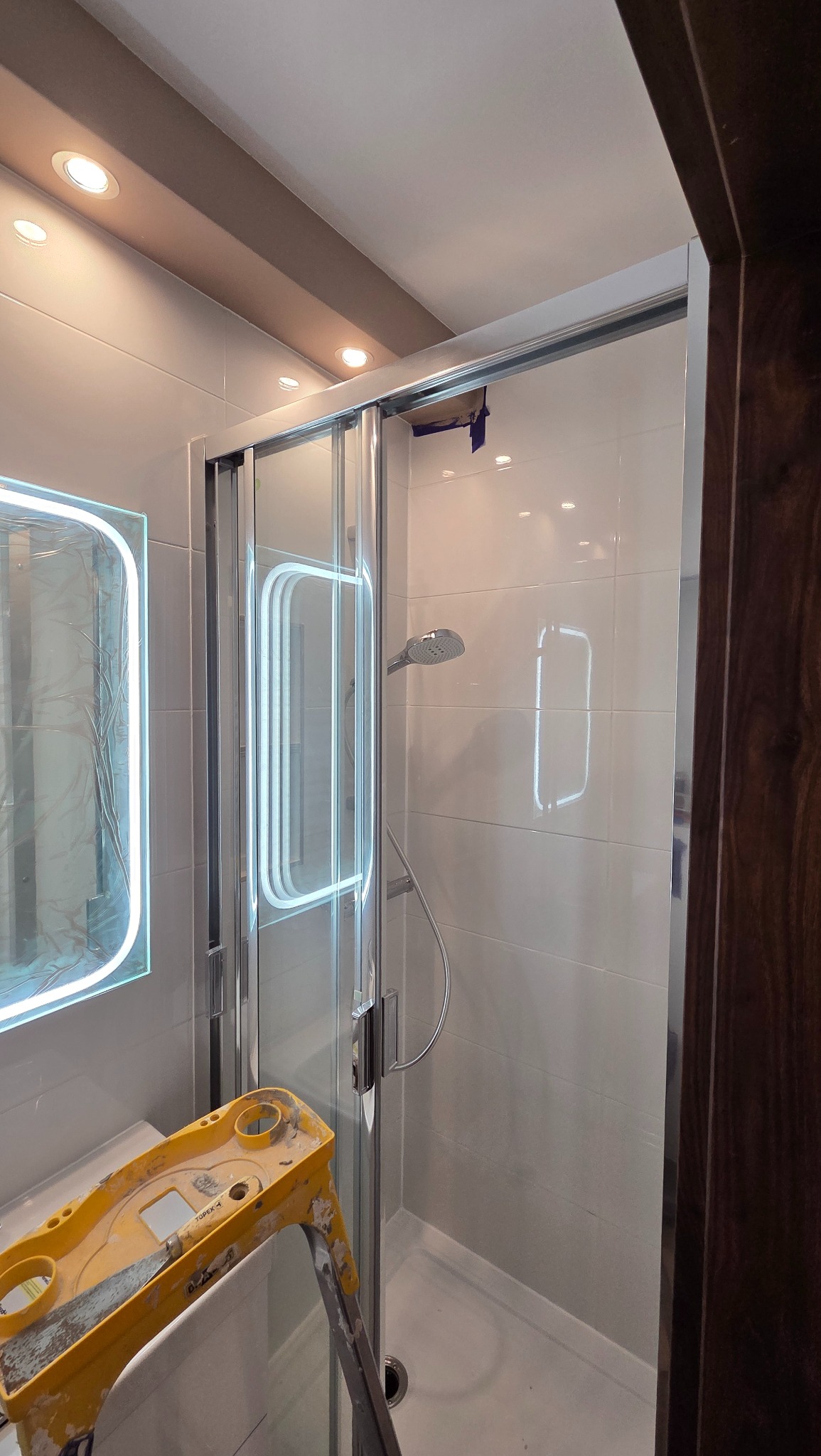 Nowoczesna kabina prysznicowa z przesuwnymi drzwiami, wykończona białymi płytkami. Widoczny fragment drabiny malarskiej i lustro z podświetleniem LED.